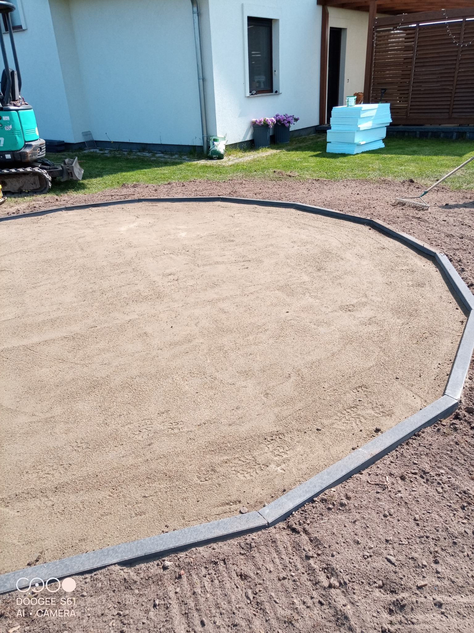 Przygotowany teren pod budowę, otoczony szarymi obrzeżami, z widocznym minikoparką w tle i stosem styropianu obok domu.