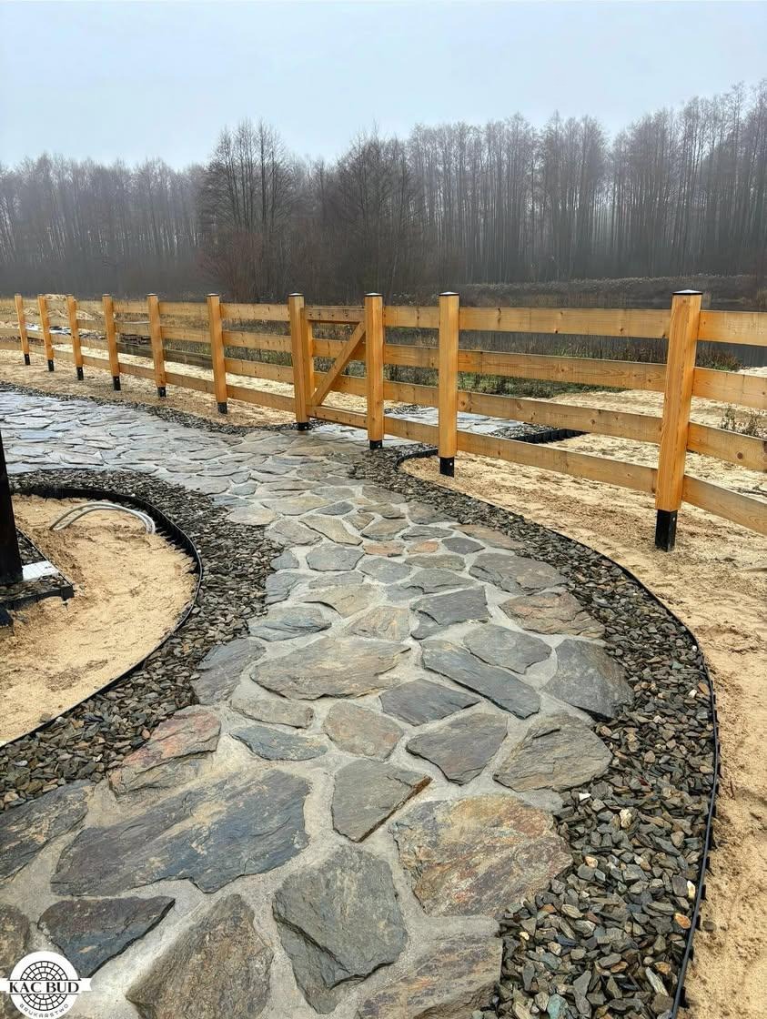 Ścieżka z nieregularnych kamieni prowadząca wzdłuż drewnianego płotu, otoczona drobnym żwirem, z lasem w tle.