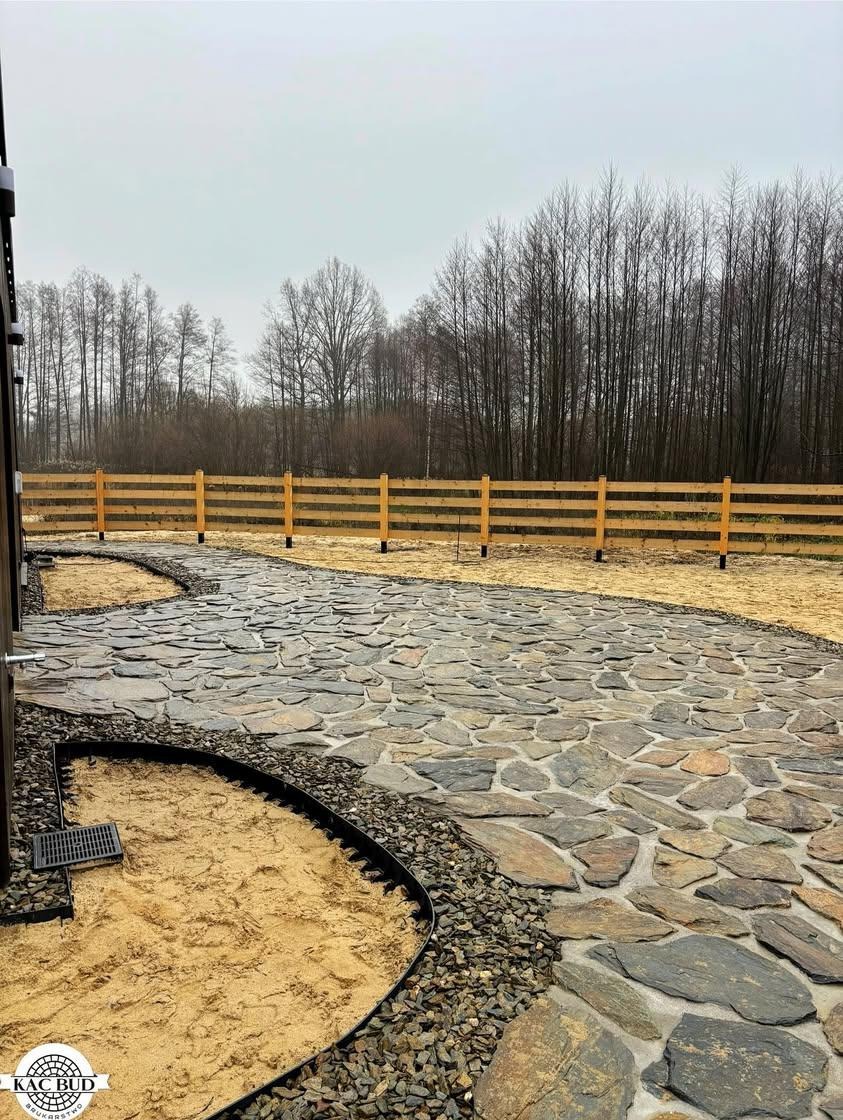 Ścieżka z nieregularnych kamiennych płyt, obrzeża z tworzywa, drewniane ogrodzenie w tle, przygotowane podłoże z piasku i żwiru.
