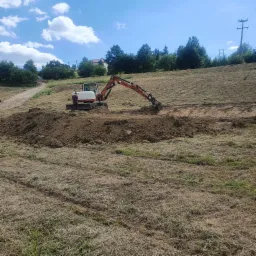 Pomarańczowa minikoparka Terex podczas wykopu w ziemi na zboczu, widoczny operator i świeżo usunięta warstwa gleby.