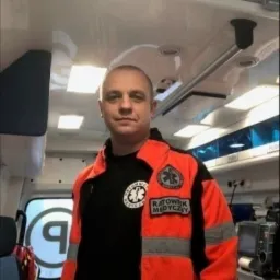 Ratownik medyczny w pomarańczowo-czarnej kurtce wewnątrz ambulansu, widoczny emblemat z napisem 'Ratownik Medyczny'.