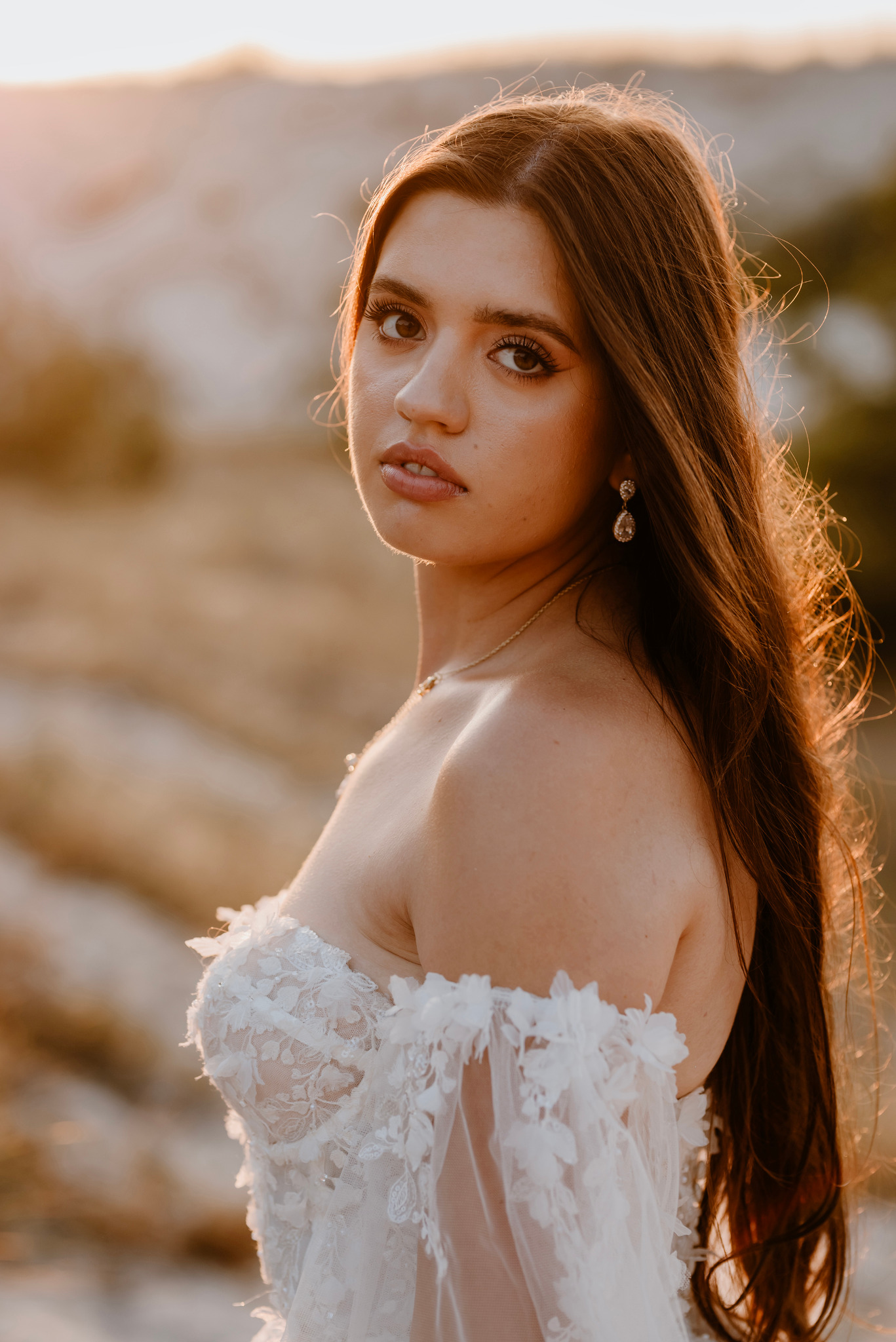 Portret kobiety w białej sukni z koronkowymi aplikacjami i długimi brązowymi włosami, pozującej na tle piaszczystych wzgórz w ciepłym świetle zachodzącego słońca.