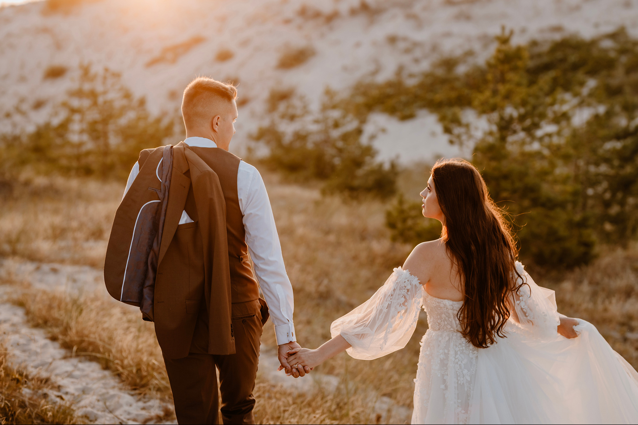 Para młoda trzymająca się za ręce na tle piaszczystych wydm o zachodzie słońca. Mężczyzna w brązowym garniturze z marynarką przewieszoną przez ramię, kobieta w białej sukni z koronkowymi rękawami.