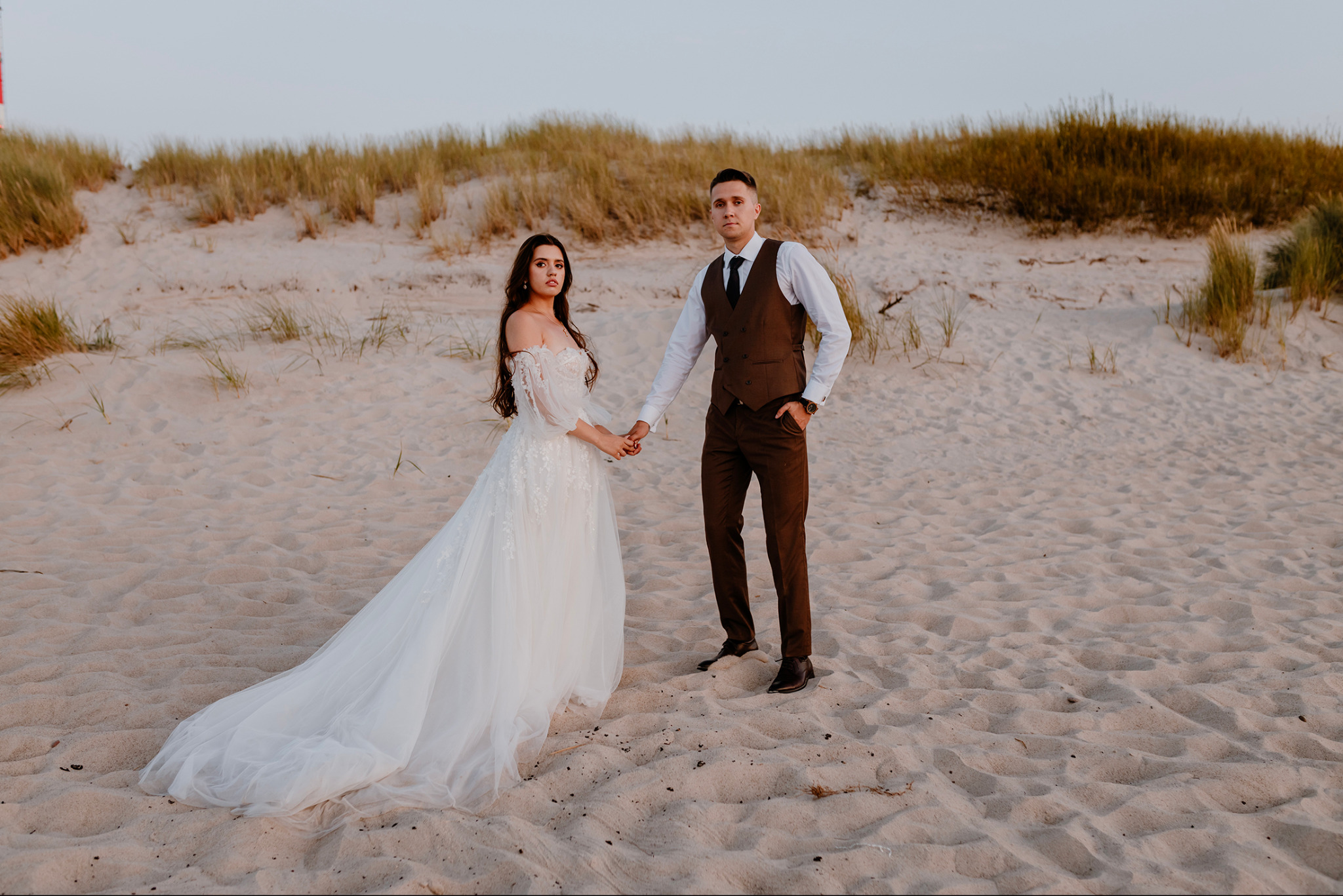 Para młoda trzymająca się za ręce na piaszczystej plaży z wydmami w tle, panna młoda w białej sukni z odkrytymi ramionami, pan młody w brązowym garniturze i czarnym krawacie.
