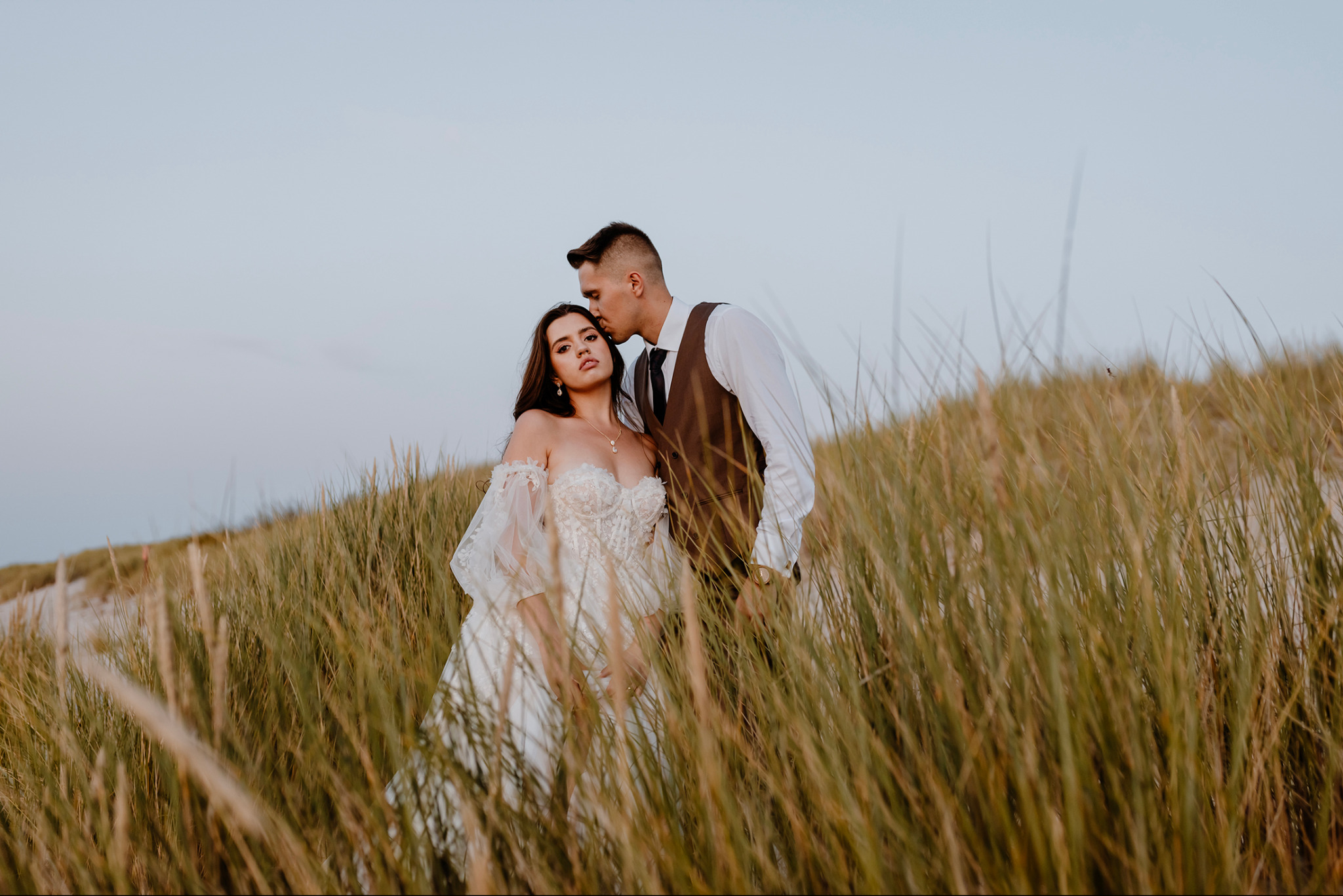 Para młoda w trawie: Panna młoda w koronkowej sukni, pan młody całuje ją w głowę, stonowana kolorystyka, naturalne światło.