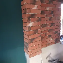Fragment ściany wykończonej czerwoną cegłą klinkierową z ciemnymi przebarwieniami, sąsiadujący z pomalowaną na ciemnozielono ścianą. Widoczne niedokończone prace wykończeniowe i narzędzia.
