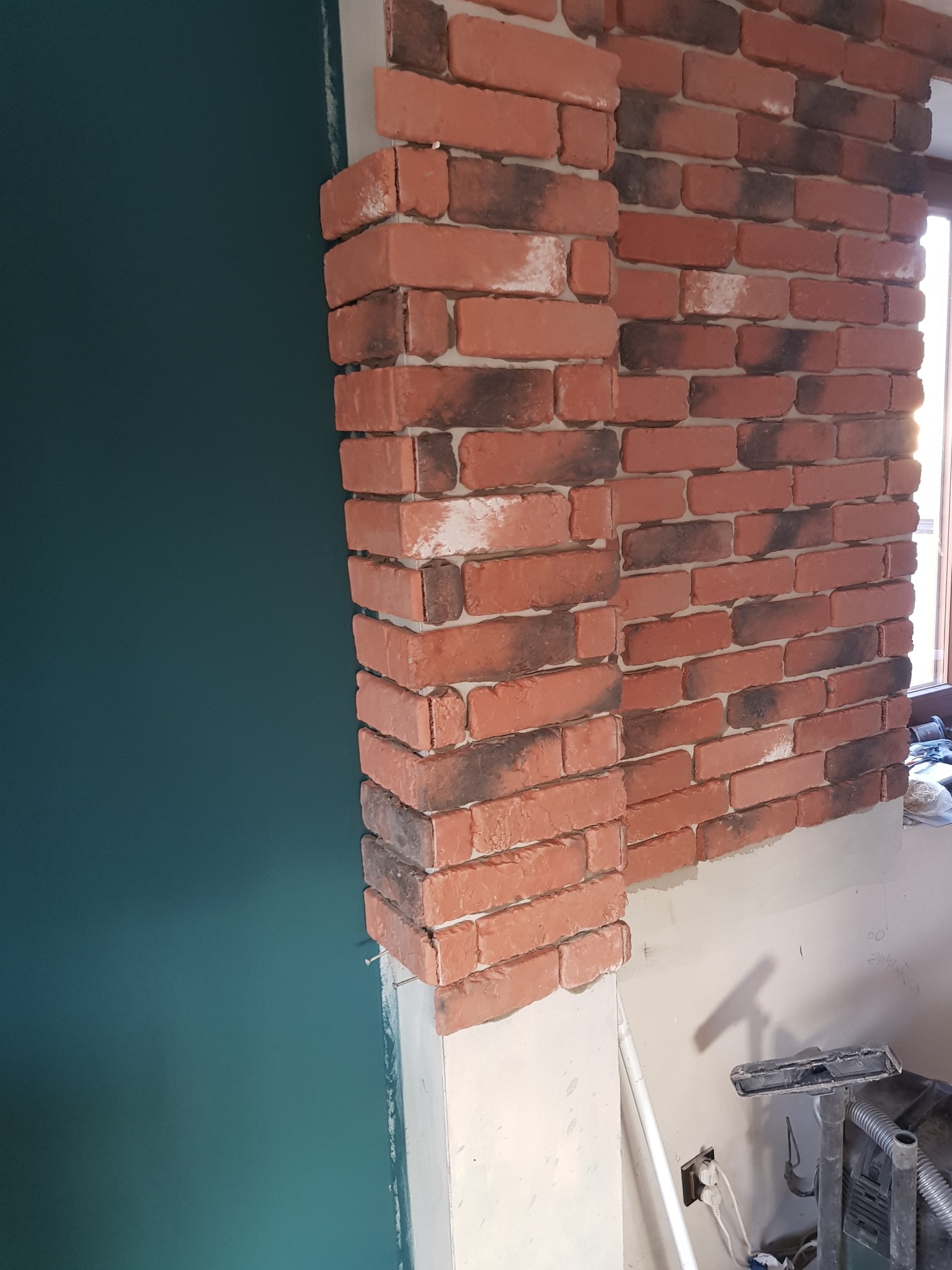 Fragment ściany wykończonej czerwoną cegłą klinkierową z ciemnymi przebarwieniami, sąsiadujący z pomalowaną na ciemnozielono ścianą. Widoczne niedokończone prace wykończeniowe i narzędzia.