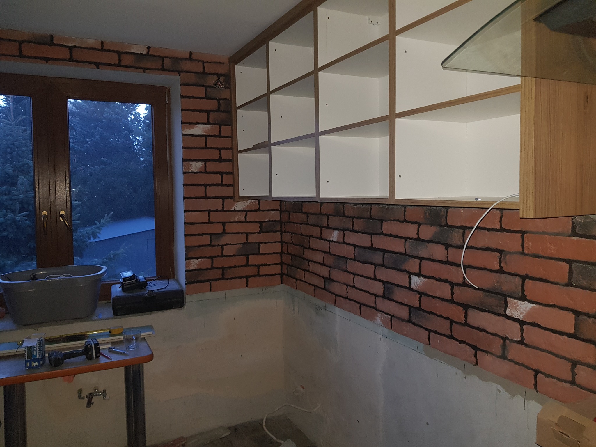 Wnętrze kuchni w trakcie prac wykończeniowych: ściana wyłożona cegłą, zamontowane białe szafki z drewnianymi ramami, widoczne narzędzia i akcesoria na stole przy oknie.