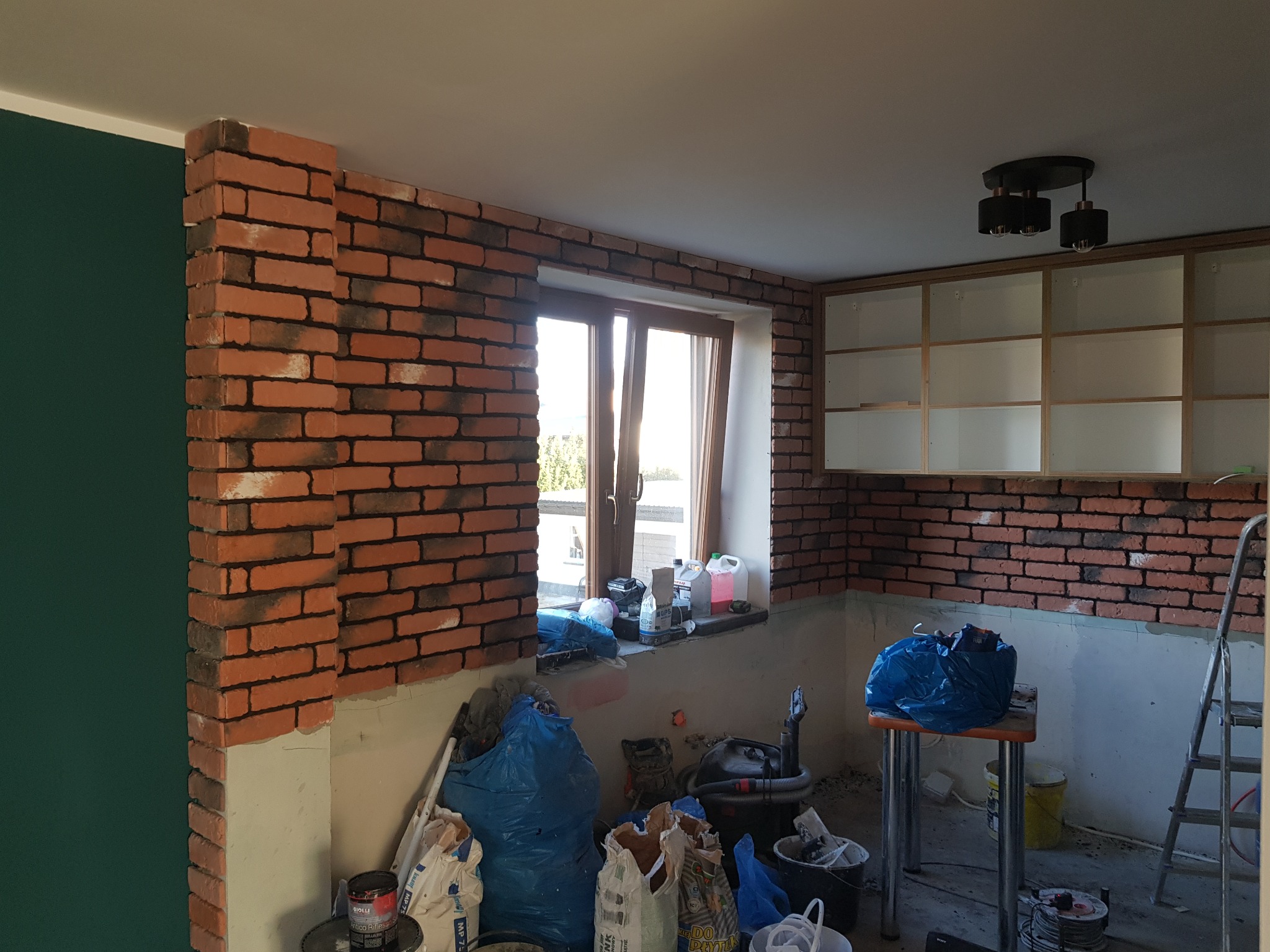 Wnętrze w trakcie prac wykończeniowych: ściany częściowo wyłożone dekoracyjną cegłą, widoczne worki z zaprawą, narzędzia i drabina, surowe szafki kuchenne bez frontów.
