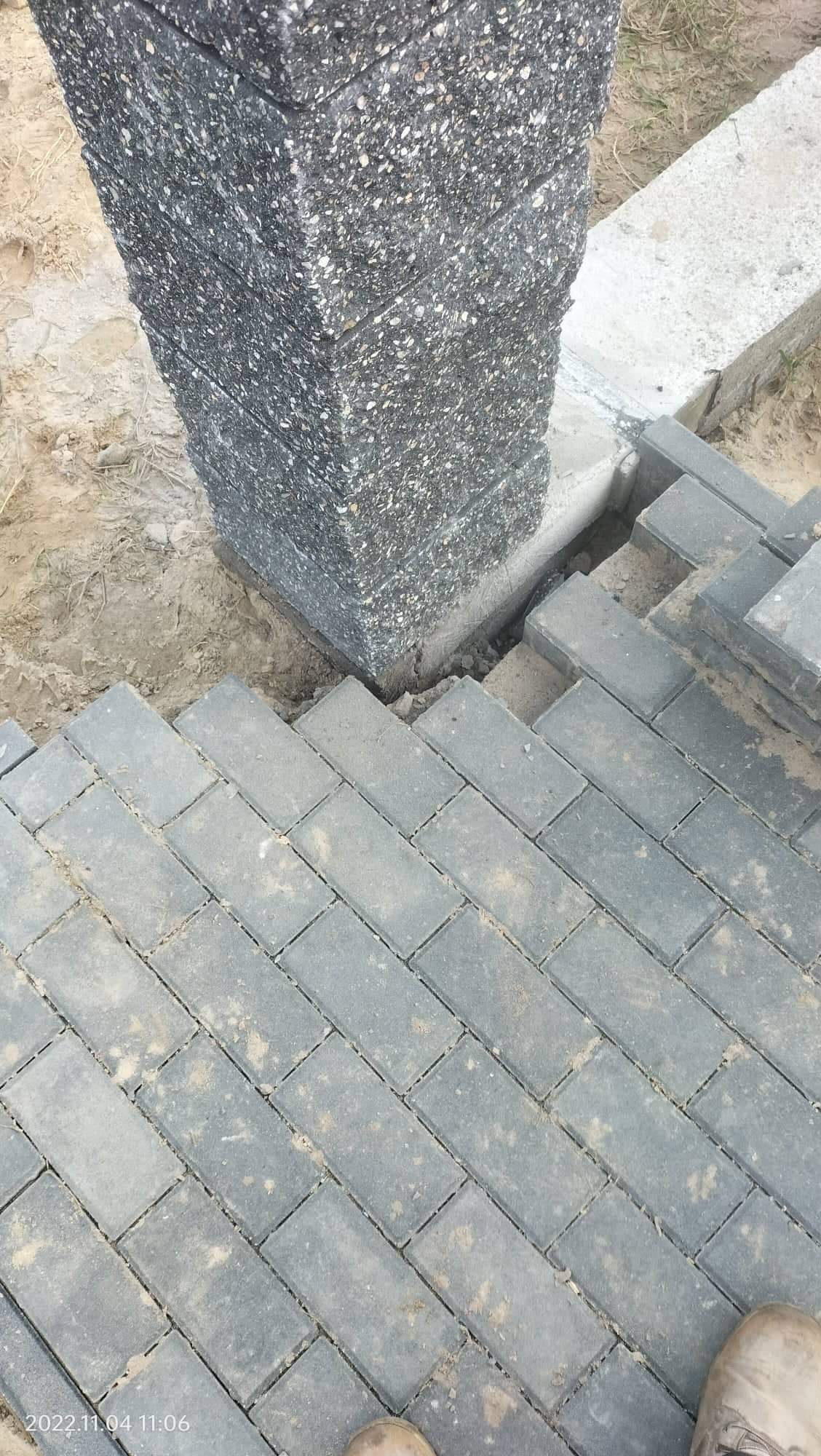 Ułożona szara kostka brukowa wokół betonowego słupka z szarego kruszywa, widoczne niedociągnięcia wokół słupka.