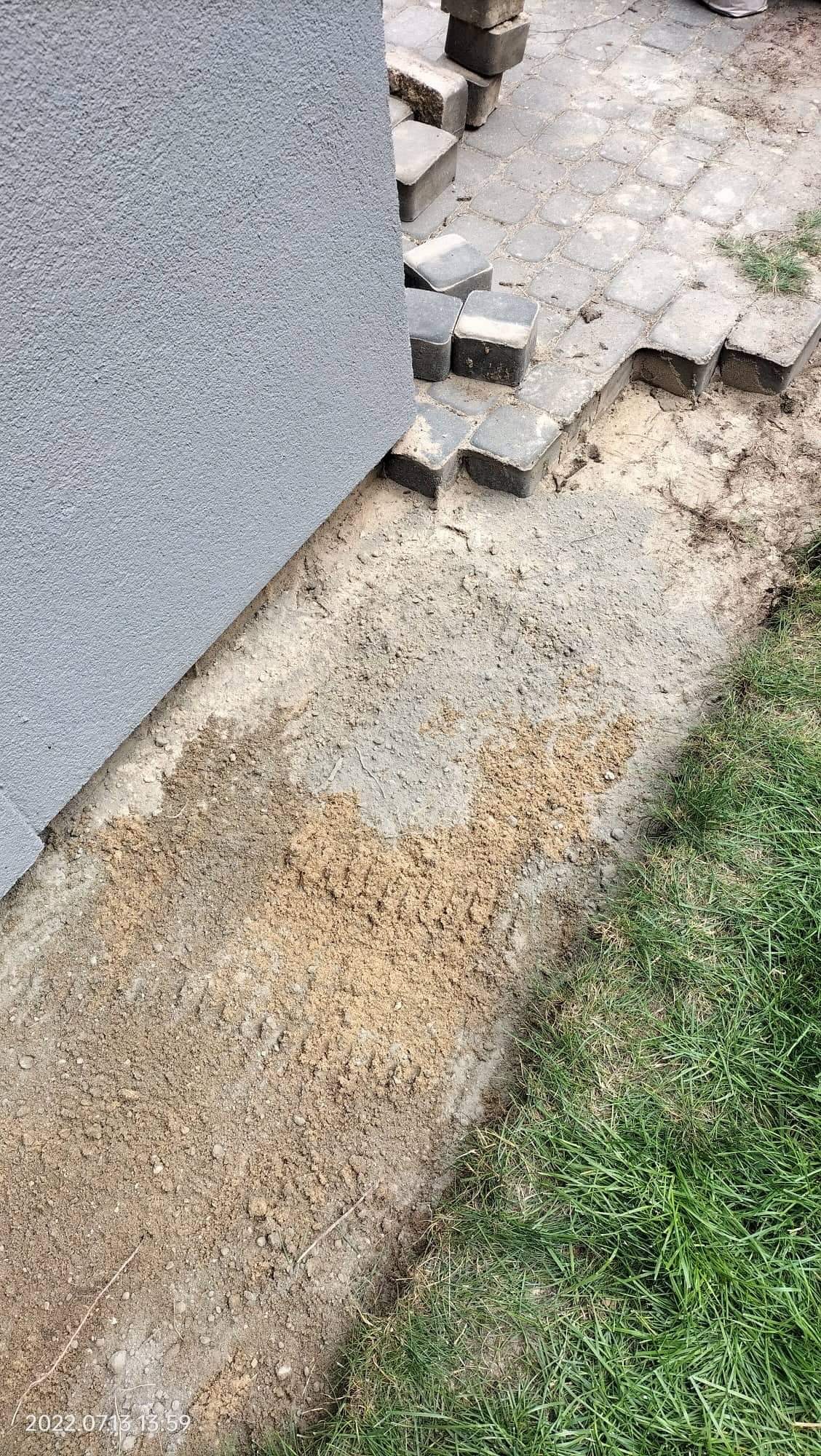 Przygotowanie podłoża pod układanie kostki brukowej przy ścianie budynku; widoczne podsypka piaskowa, częściowo ułożona kostka i trawa.