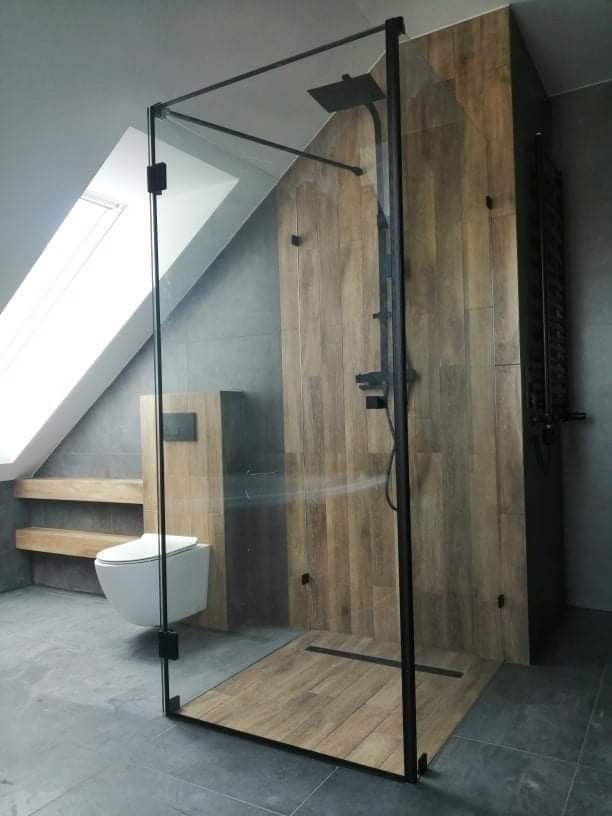 Nowoczesna kabina prysznicowa typu walk-in z czarnymi profilami, drewnopodobne płytki na ścianie i podłodze, wisząca toaleta z podtynkowym systemem spłukiwania w łazience na poddaszu.
