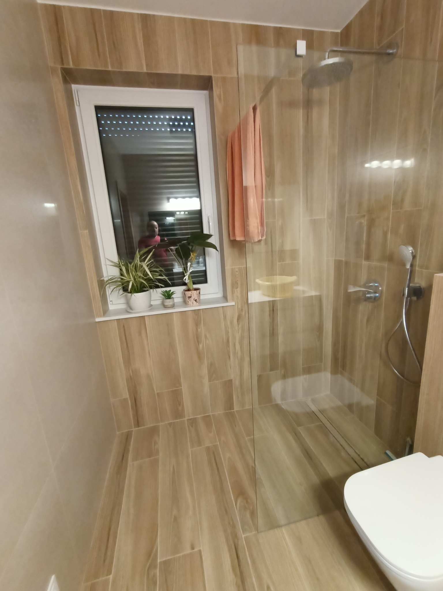 Jasna łazienka z kabiną prysznicową typu walk-in, wykończona płytkami imitującymi drewno, z oknem ozdobionym roślinami doniczkowymi i widocznym odbiciem osoby w szybie.