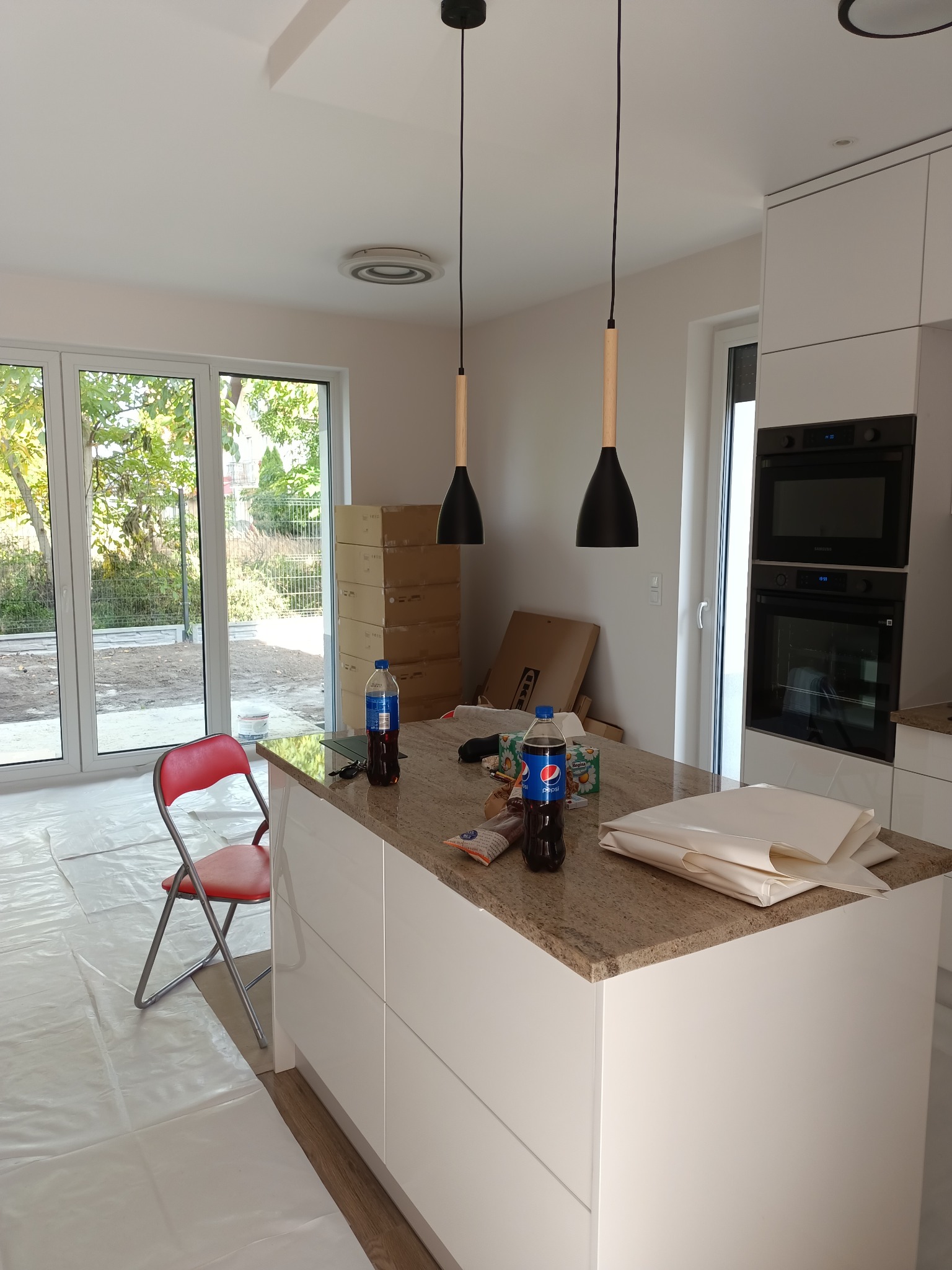 Wnętrze jasnej kuchni w trakcie wykańczania, z wyspą z blatem imitującym kamień, podwieszanymi lampami z czarnymi kloszami i drewnianymi elementami, podłoga zabezpieczona folią ochronną, w tle...