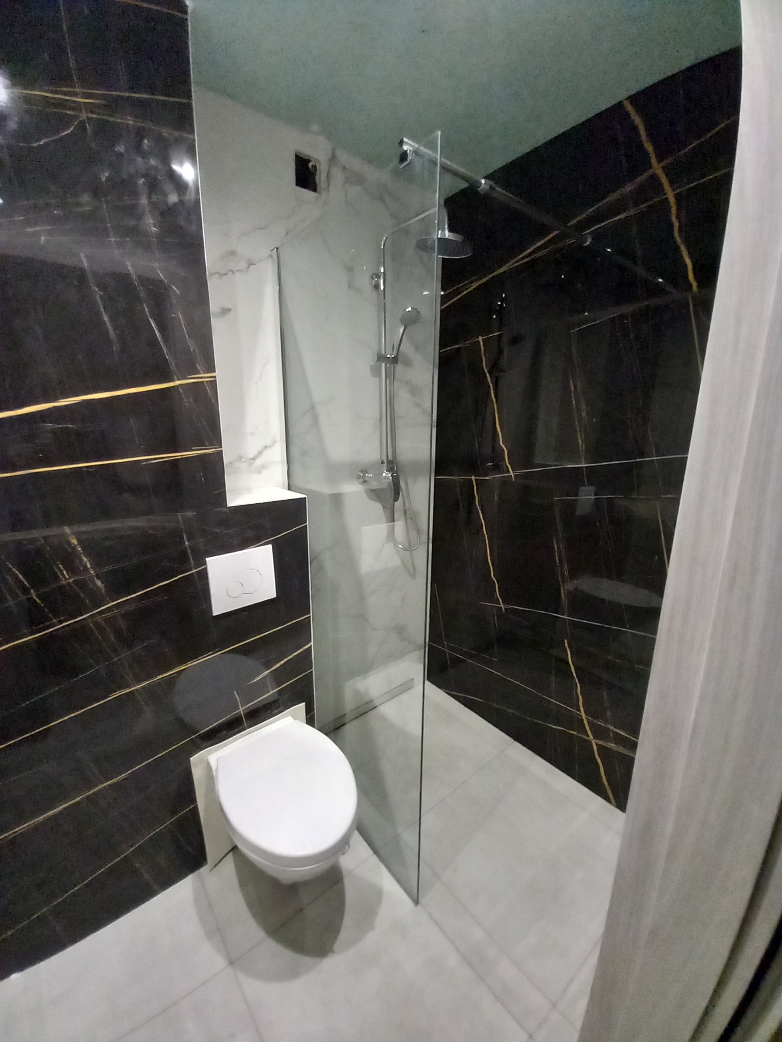 Nowoczesna łazienka z czarnymi płytkami imitującymi marmur z złotymi żyłami, białą ceramiką sanitarną i kabiną prysznicową ze szklaną ścianką.