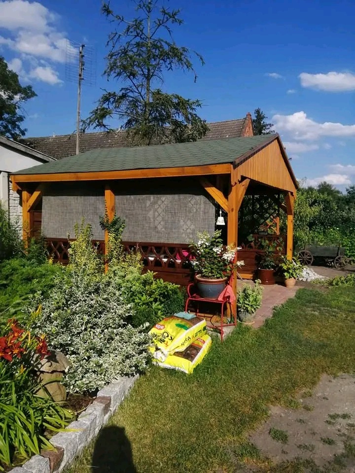 Drewniana altana z zielonym dachem w ogrodzie, z zasłoniętą jedną ścianą, obok rośliny i worki ziemi.