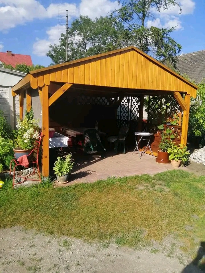 Drewniana altana ogrodowa z meblami, otoczona zielenią i kwiatami w donicach, wyłożona kostką brukową.