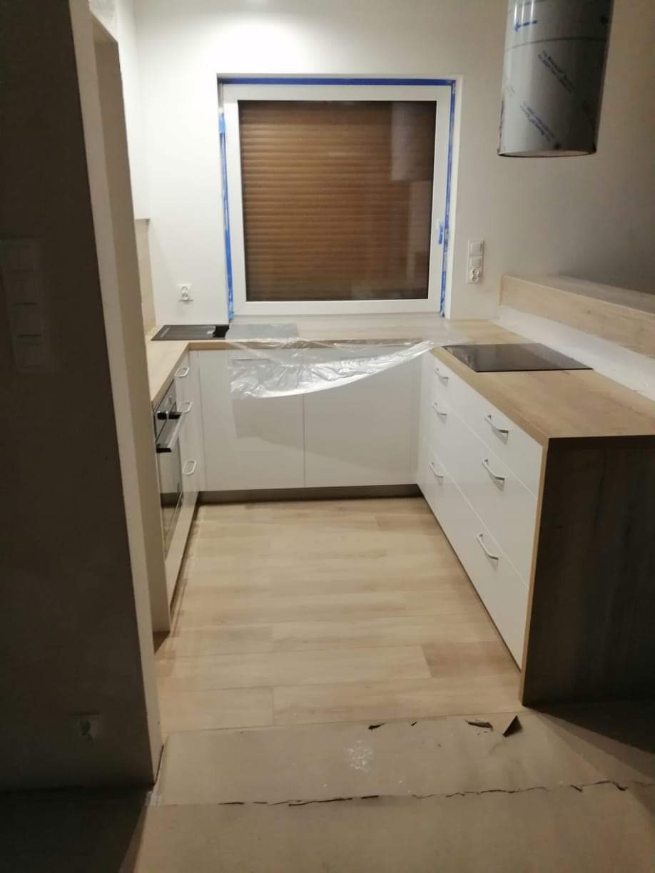 Nowoczesna kuchnia w trakcie wykończenia: białe szafki z drewnianym blatem, częściowo zabezpieczone folią, okno z brązowymi żaluzjami, jasna podłoga imitująca drewno, widoczny fragment okapu...