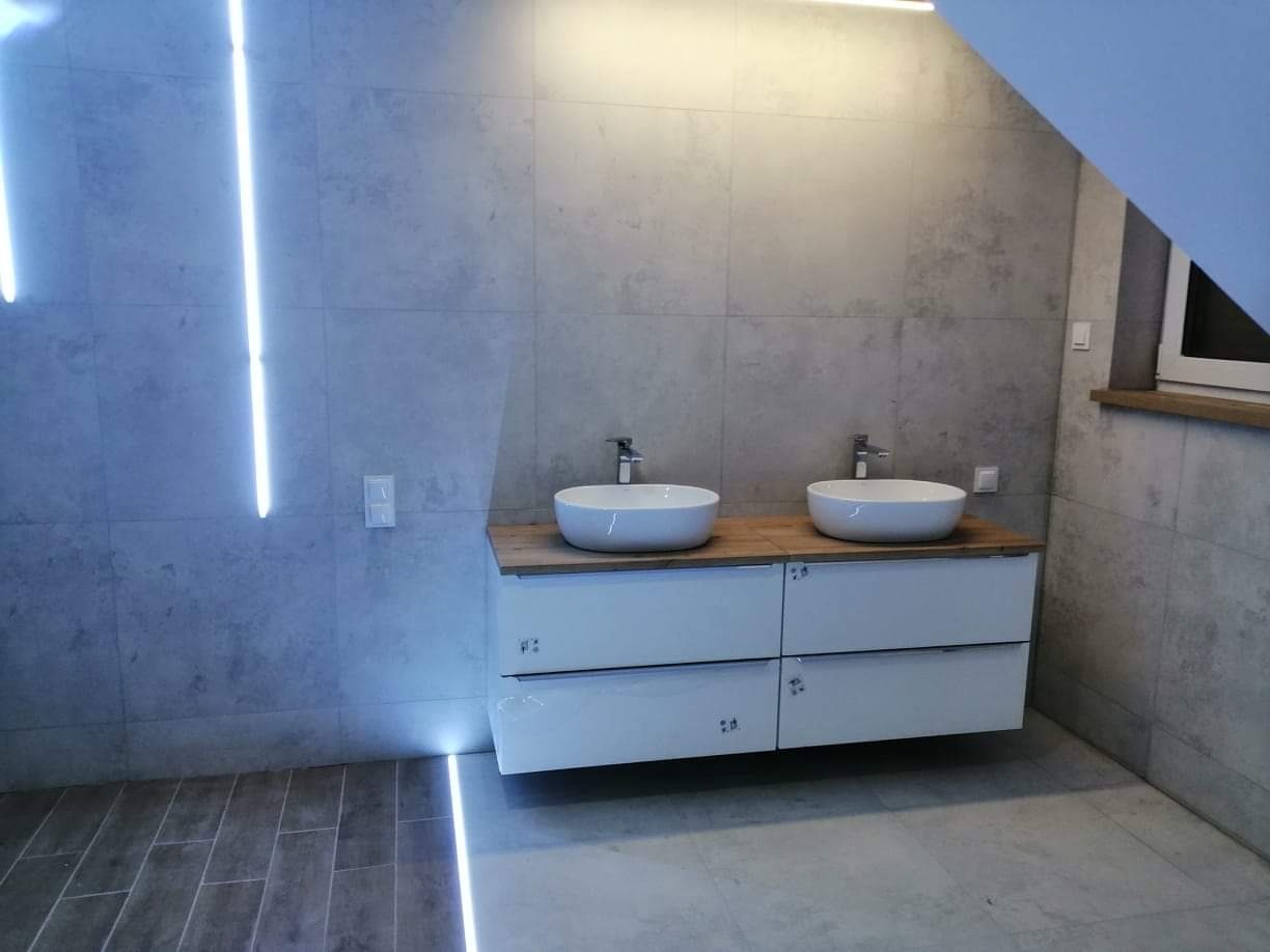 Nowoczesna łazienka z dwoma okrągłymi umywalkami na drewnianym blacie, białe szafki podwieszane, szare płytki ścienne i podłogowe, oświetlenie LED w pionowych pasach i pod szafkami, okno w skosie...