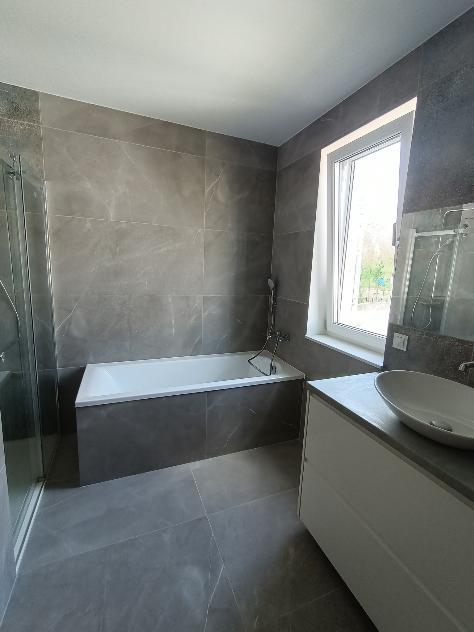Nowoczesna łazienka w odcieniach szarości z marmurkowymi płytkami, białą wanną i umywalką nablatową na szafce, widok z kabiny prysznicowej.