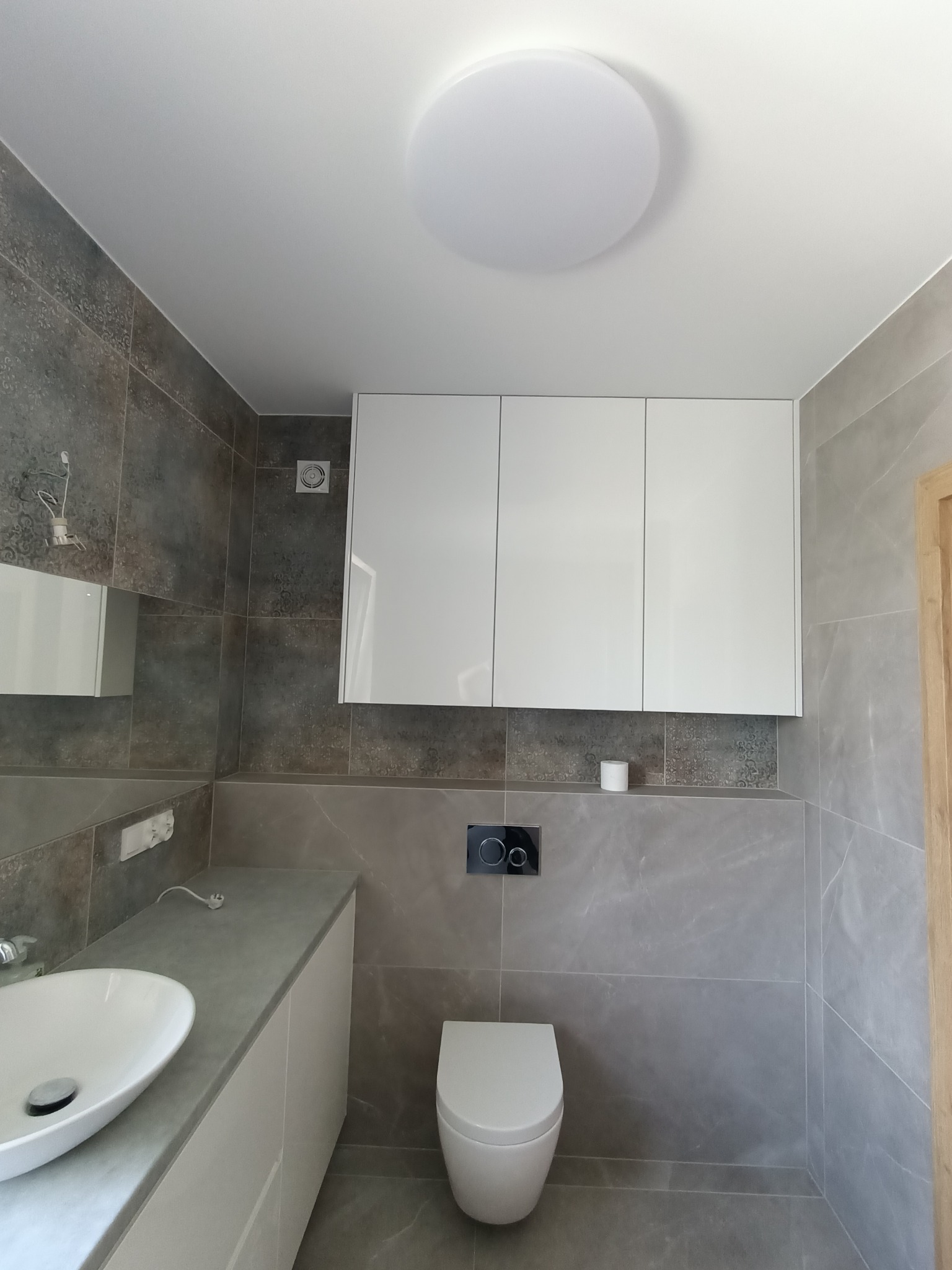 Nowoczesna łazienka z szarymi płytkami imitującymi kamień, białą umywalką nablatową, podwieszaną miską WC i szafkami wiszącymi pod sufitem z okrągłym plafonem.