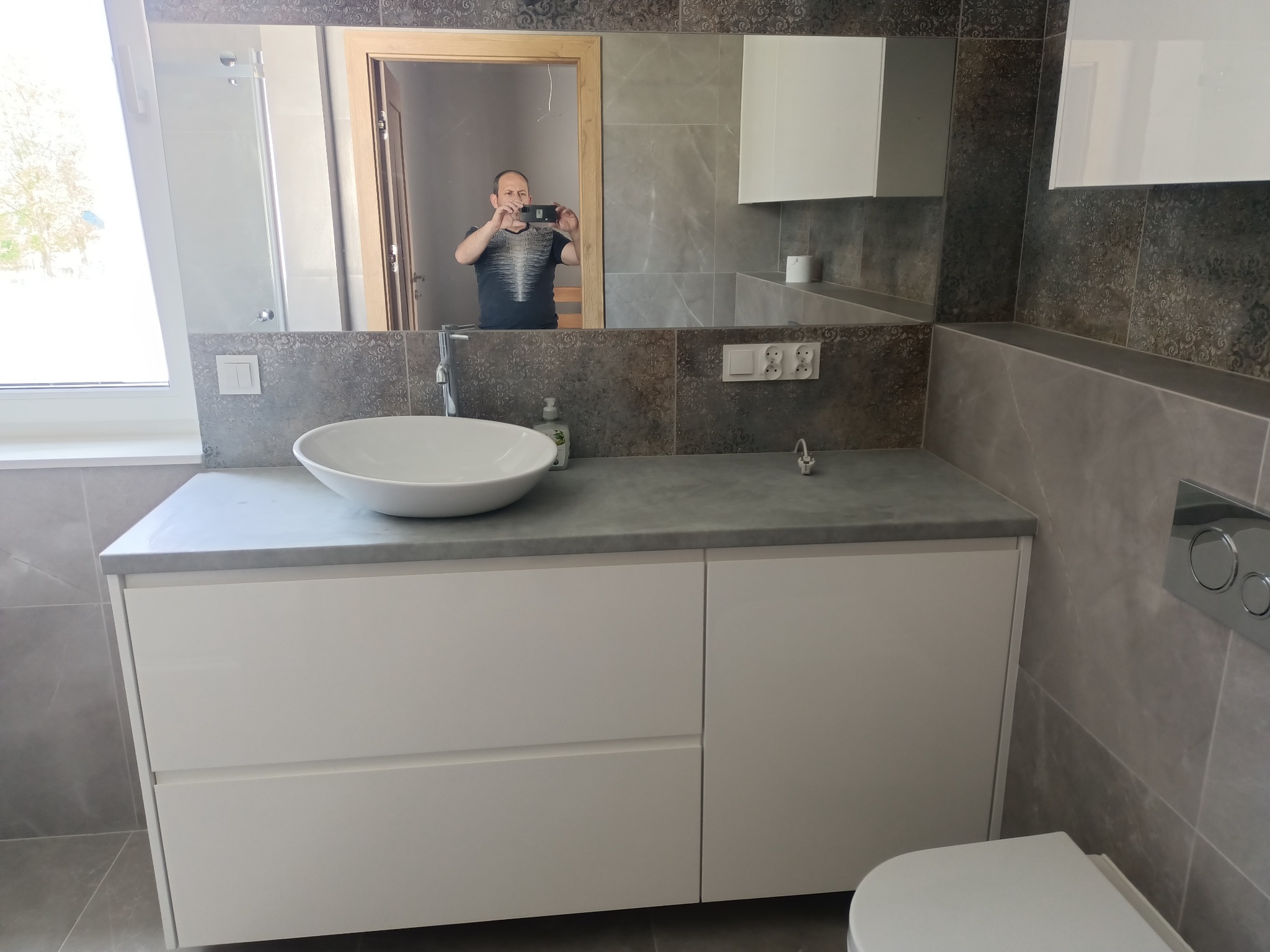 Nowoczesna łazienka z szarymi płytkami, białą umywalką nablatową i szafką pod umywalką. W lustrze odbija się osoba robiąca zdjęcie telefonem.