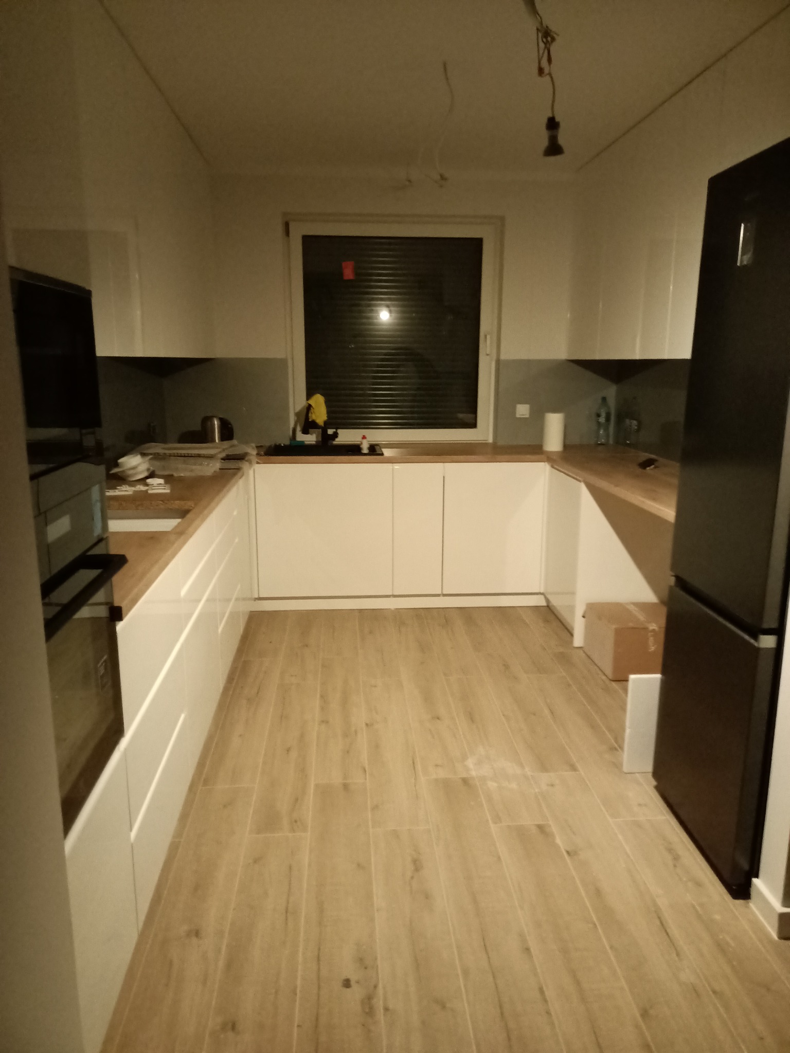 Ujęcie wnętrza kuchni z białymi, lakierowanymi szafkami, drewnianym blatem i podłogą imitującą drewno, widoczne niedokończone podłączenie oświetlenia sufitowego.