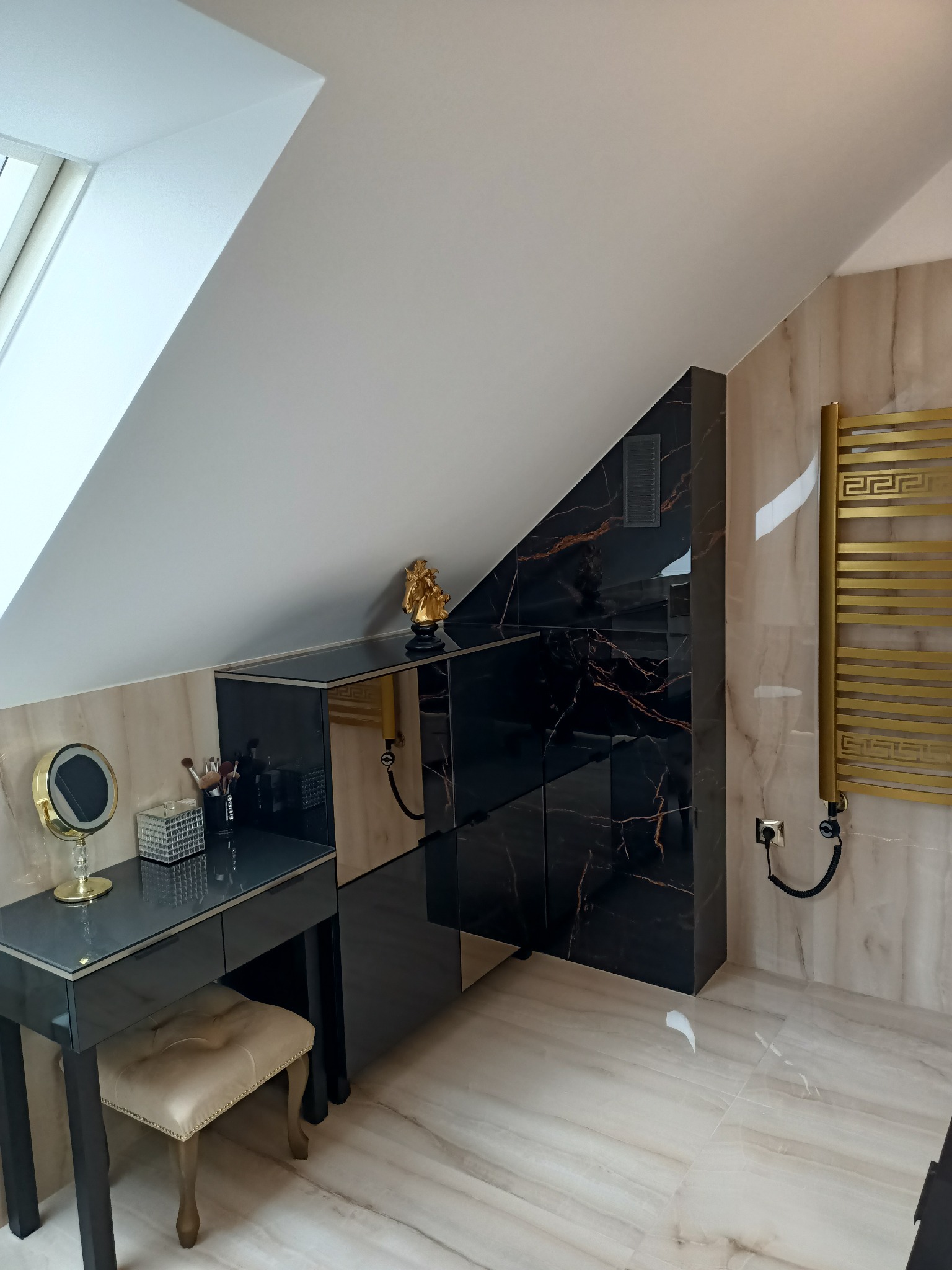 Nowoczesna łazienka na poddaszu z marmurowymi płytkami, czarną zabudową meblową z elementami złota oraz grzejnikiem drabinkowym w kolorze złotym.