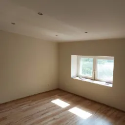 Pomalowane na beżowo puste pomieszczenie z nowo ułożonymi panelami podłogowymi w odcieniach brązu i oknem z białymi ramami.