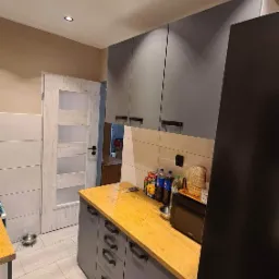 Nowoczesna kuchnia po remoncie: szare szafki z drewnianym blatem, jasne płytki podłogowe imitujące drewno, widoczny fragment lodówki i drzwi do innego pomieszczenia.