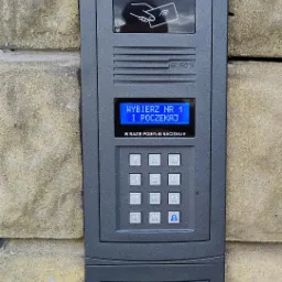 Szary panel domofonu z klawiaturą numeryczną i czytnikiem kart zbliżeniowych zamontowany na ścianie z bloków. Na ekranie wyświetla się komunikat 'Wybierz nr 1 i poczekaj'.