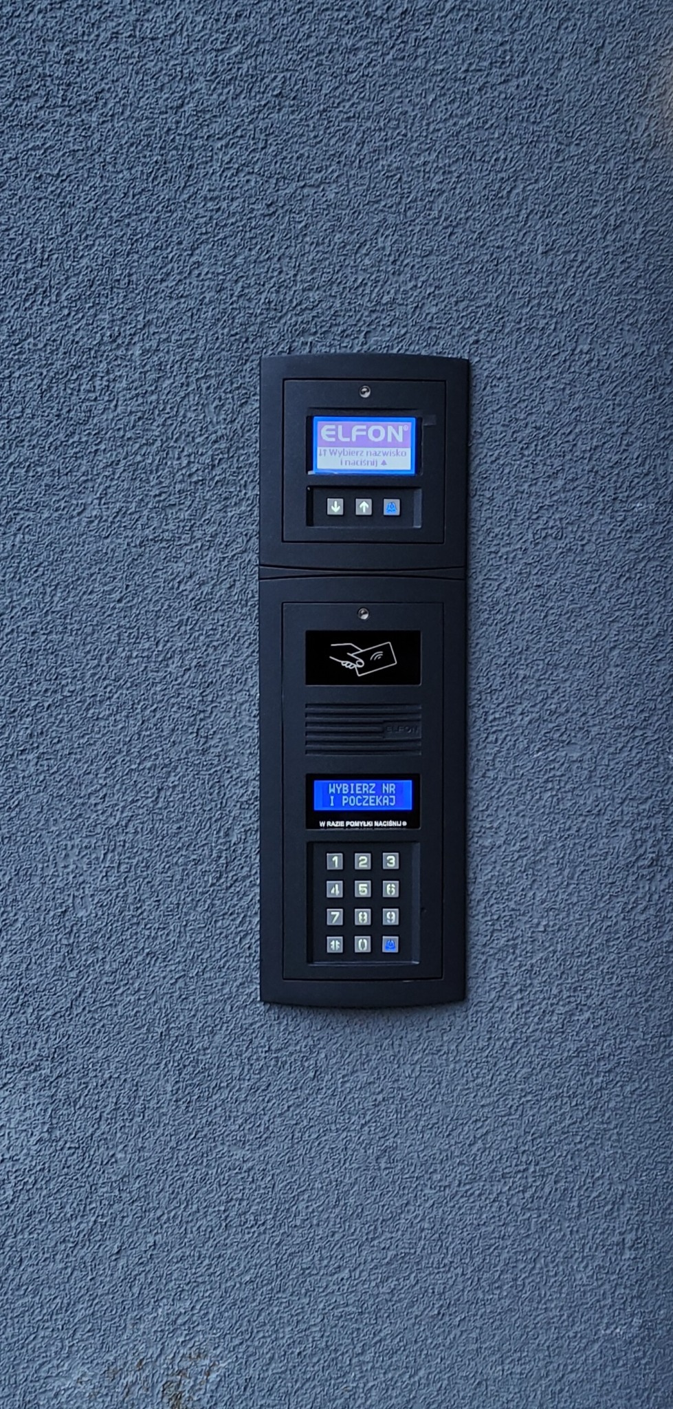 Czarny domofon Elfon z wyświetlaczem i klawiaturą numeryczną zamontowany na szarej, chropowatej ścianie zewnętrznej.