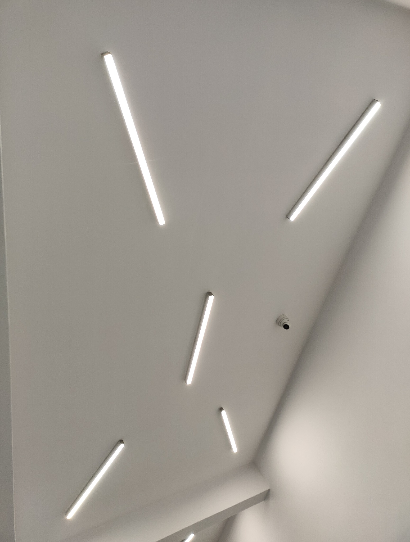 Biały sufit z rozmieszczonymi liniowo lampami LED i czujnikiem dymu, widok z dołu pod kątem.