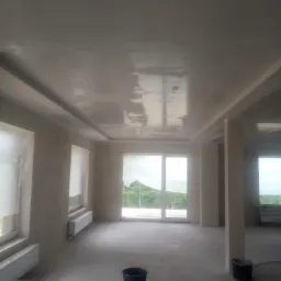 Wnętrze w trakcie prac wykończeniowych, ściany i sufit pokryte świeżą gładzią, okna zabezpieczone folią, widoczne dwa wiadra i grzejniki.