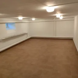 Wykończone pomieszczenie piwniczne z brązowymi płytkami podłogowymi, białymi ścianami i rurami biegnącymi wzdłuż ścian, oświetlone przez szereg okrągłych lamp sufitowych.