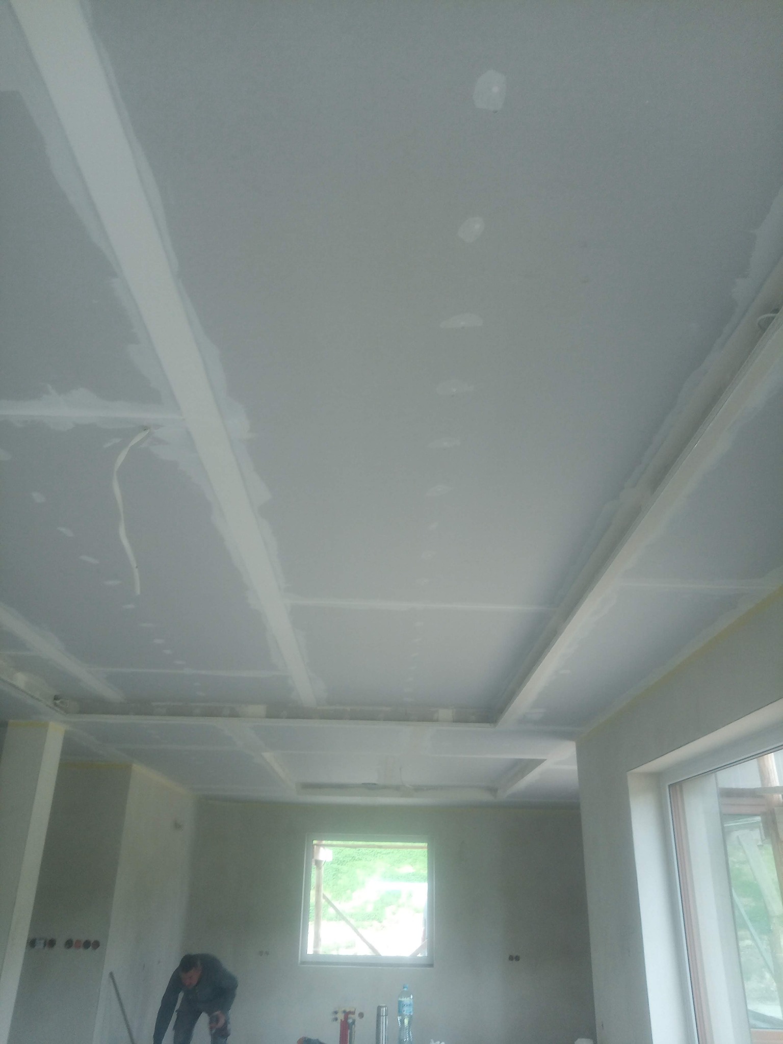 Sufit podwieszany z płyt gipsowo-kartonowych, częściowo wyszpachlowany, widoczne łączenia płyt, w tle pracownik w trakcie szpachlowania ścian.