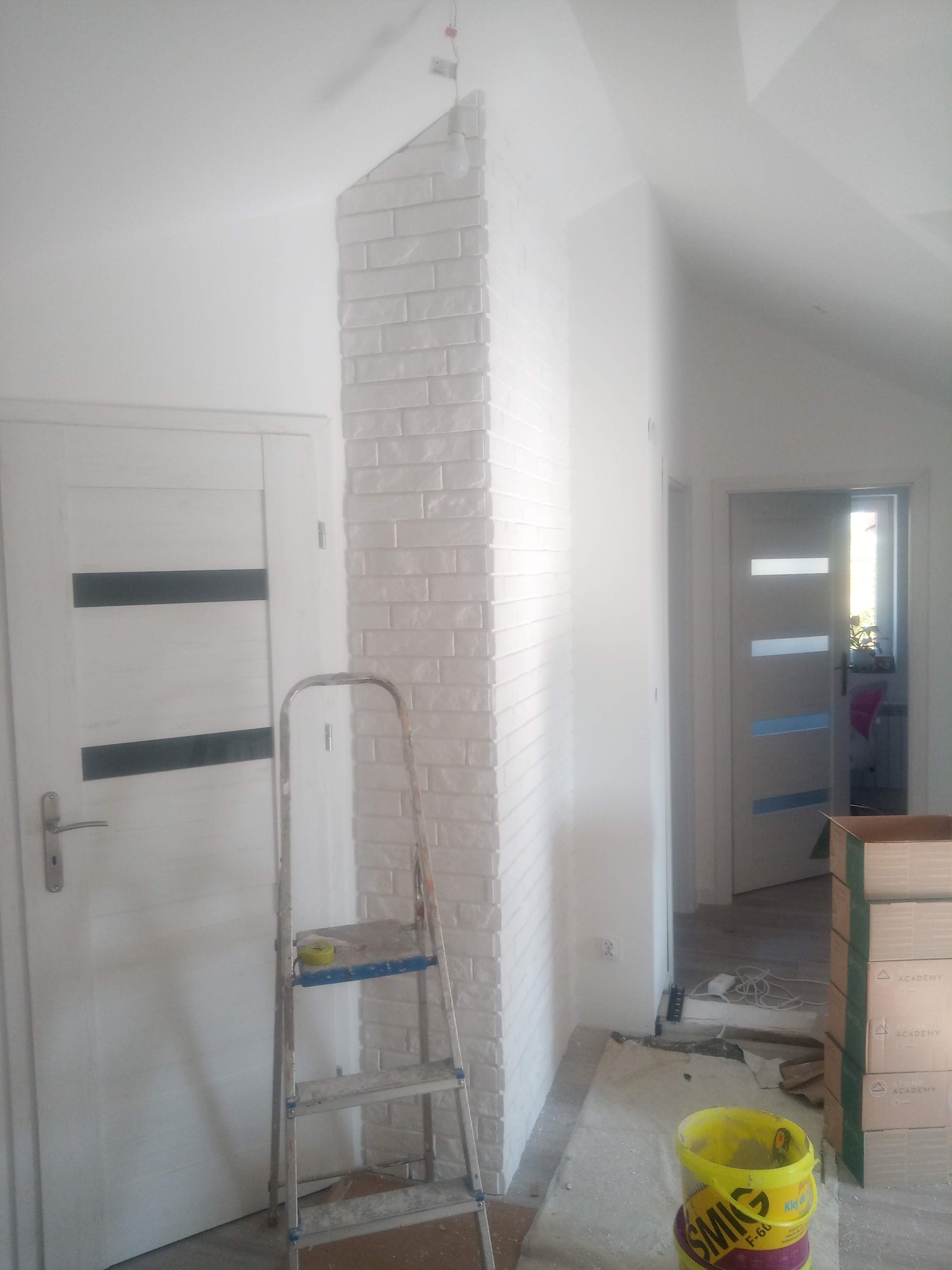 Wnętrze w trakcie prac wykończeniowych: fragment ściany wyłożony białą cegłą dekoracyjną, drabina aluminiowa z miarką, wiadro z klejem i kartony w tle, drzwi wewnętrzne z czarnymi wstawkami po...
