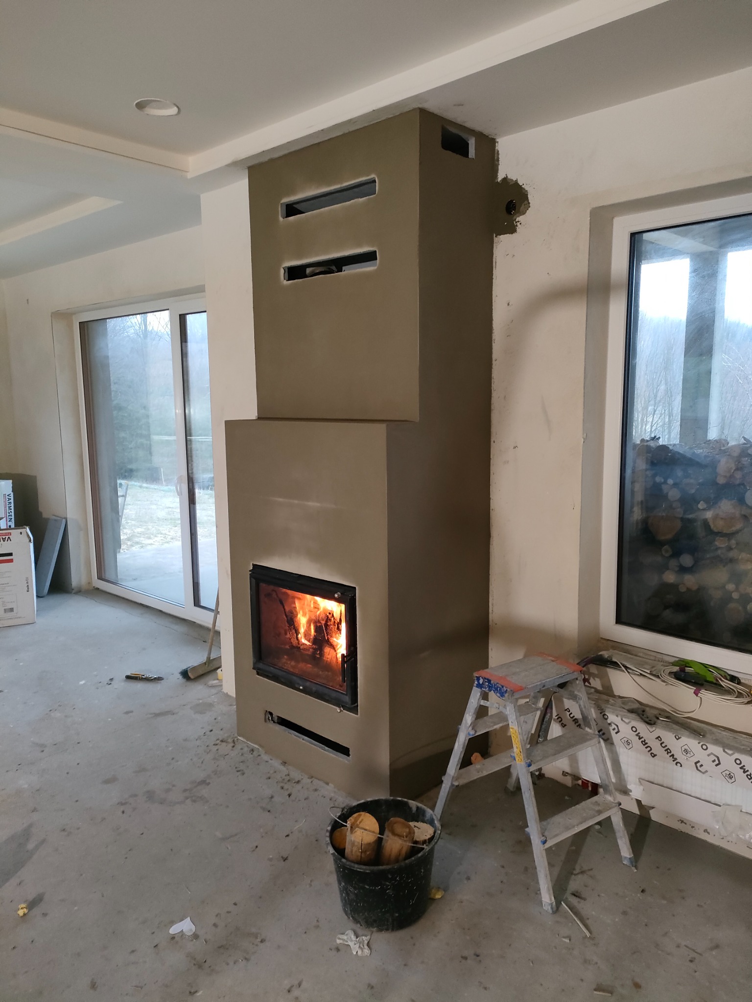 Murowany kominek z szarą gładzią w trakcie wykańczania, z palącym się ogniem wewnątrz, ustawiony w surowym wnętrzu z widocznym oknem, drabiną i wiadrem z drewnem.