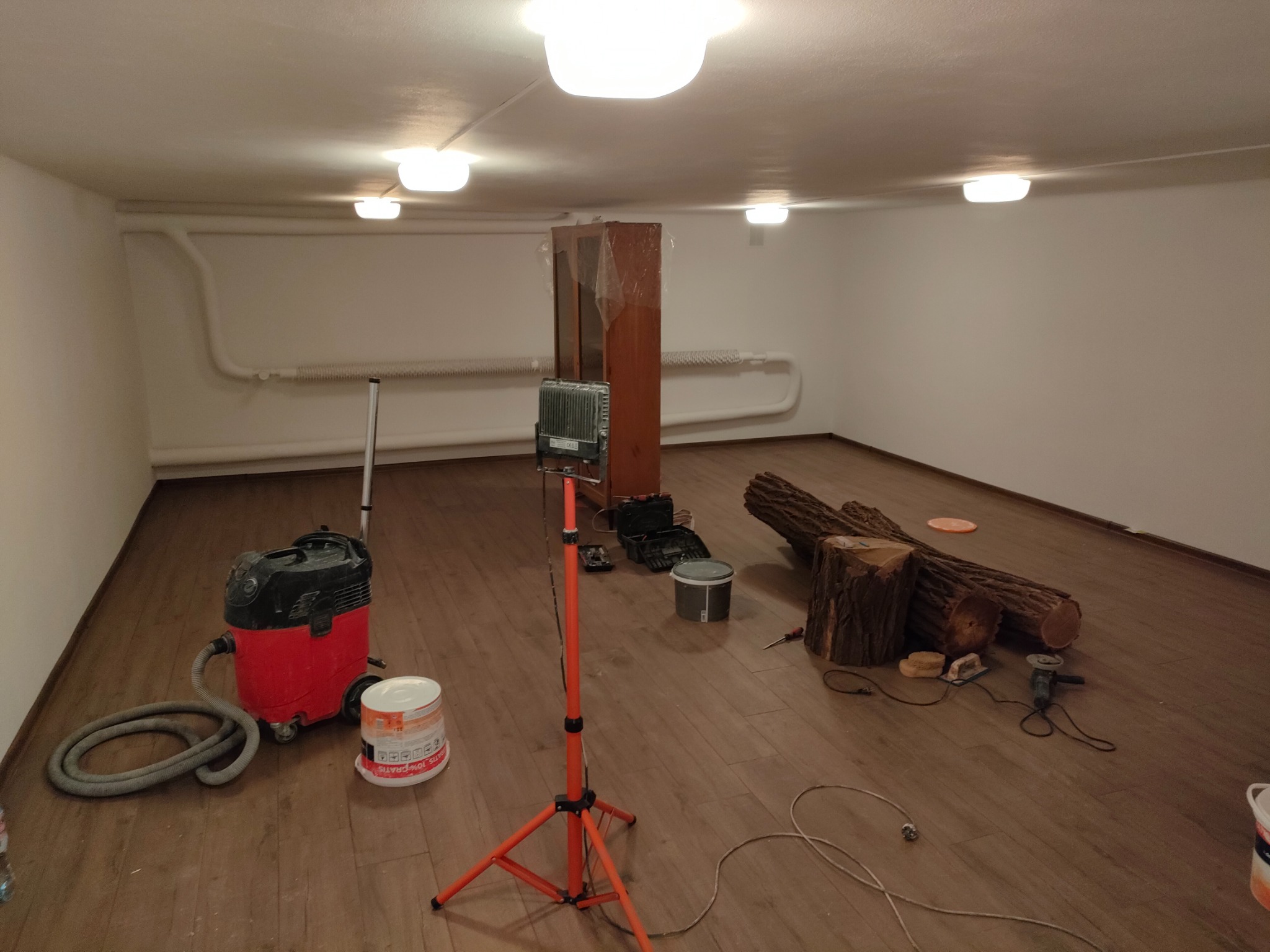 Przygotowania do prac wykończeniowych w pustym pomieszczeniu z drewnianą podłogą: odkurzacz przemysłowy, wiadro z farbą, lampa budowlana, narzędzia i pocięte pnie drzew.