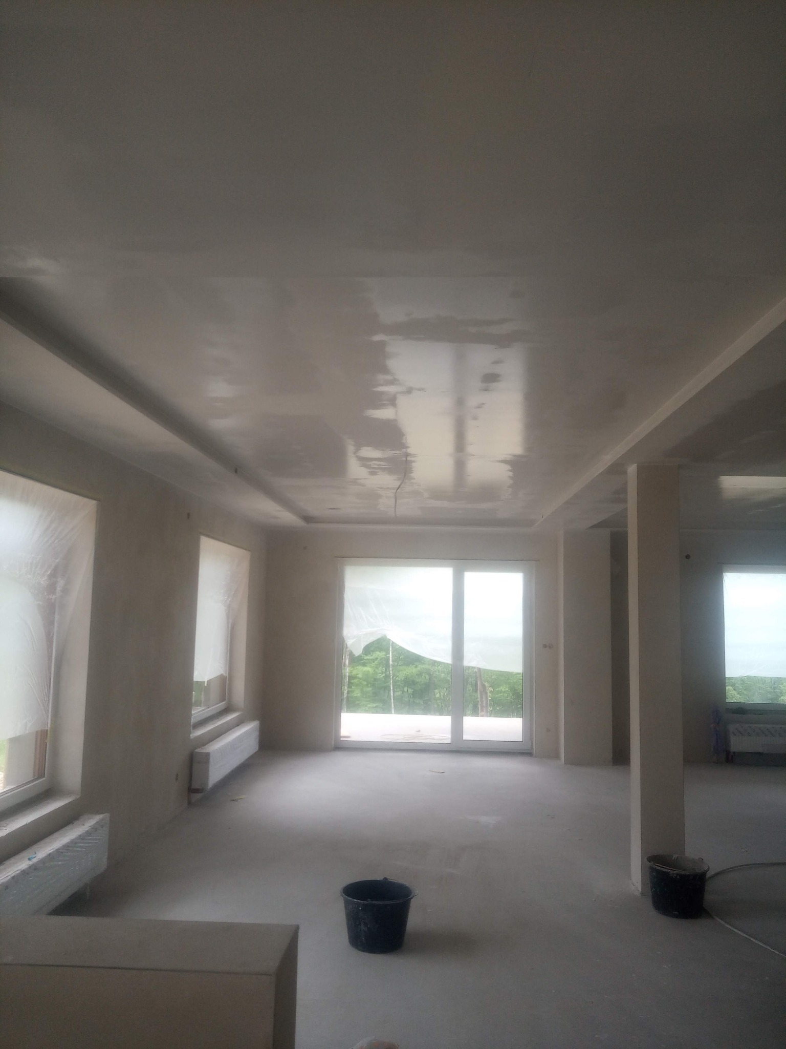 Wnętrze w trakcie prac wykończeniowych, ściany i sufit pokryte świeżą gładzią, okna zabezpieczone folią, widoczne dwa wiadra i grzejniki.