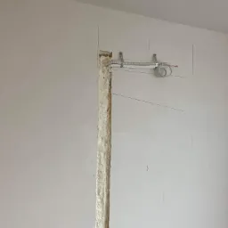 Ściana w trakcie prac instalacyjnych: pionowy wyżłobiony kanał wypełniony pianką montażową, powyżej rurka karbowana przymocowana do ściany metalowymi uchwytami, widoczne przewody elektryczne.