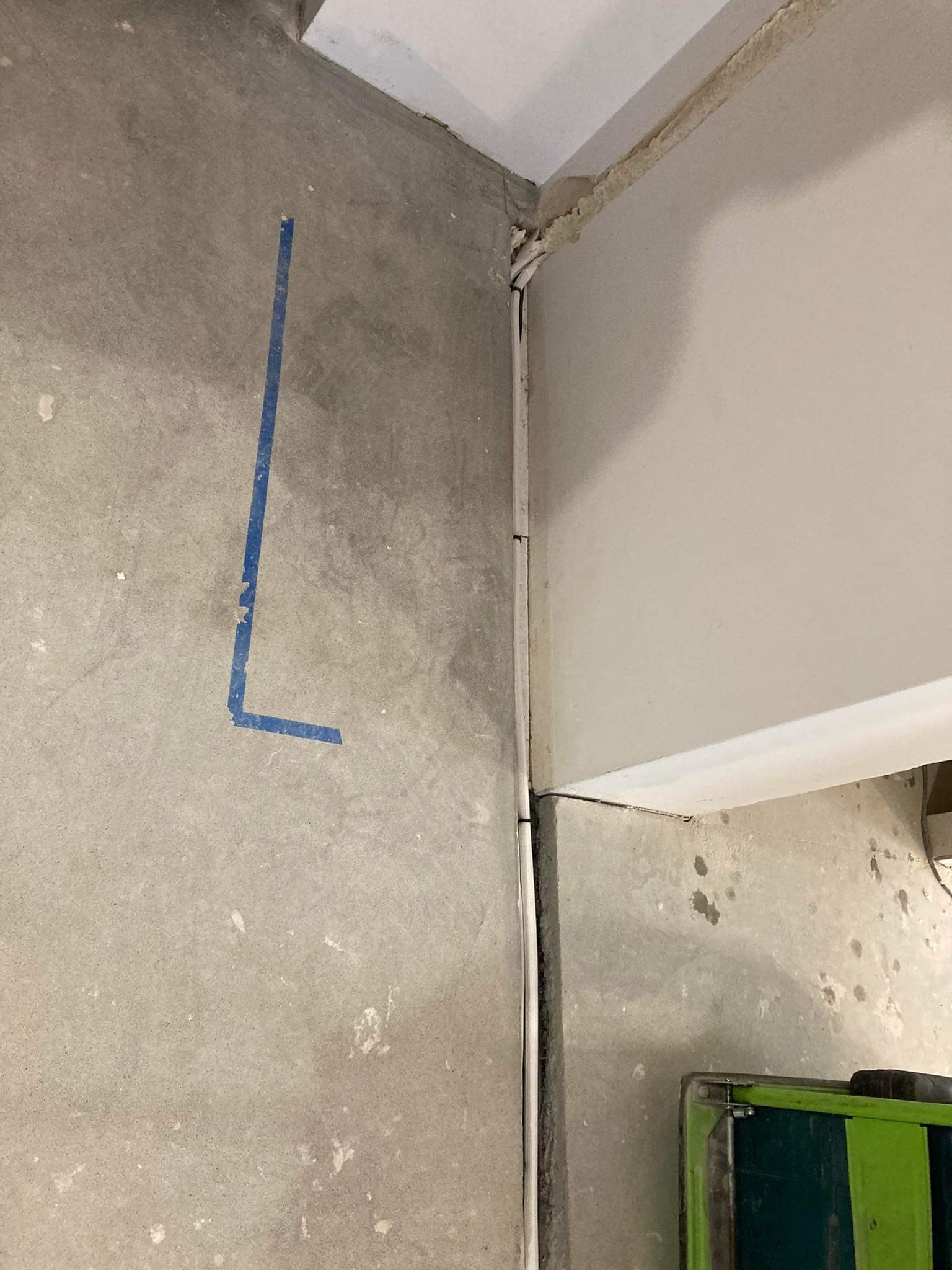 Surowa ściana w trakcie prac instalacyjnych z poprowadzoną białą listwą kablową i niebieską taśmą wyznaczającą pion, widoczny fragment skrzynki narzędziowej.