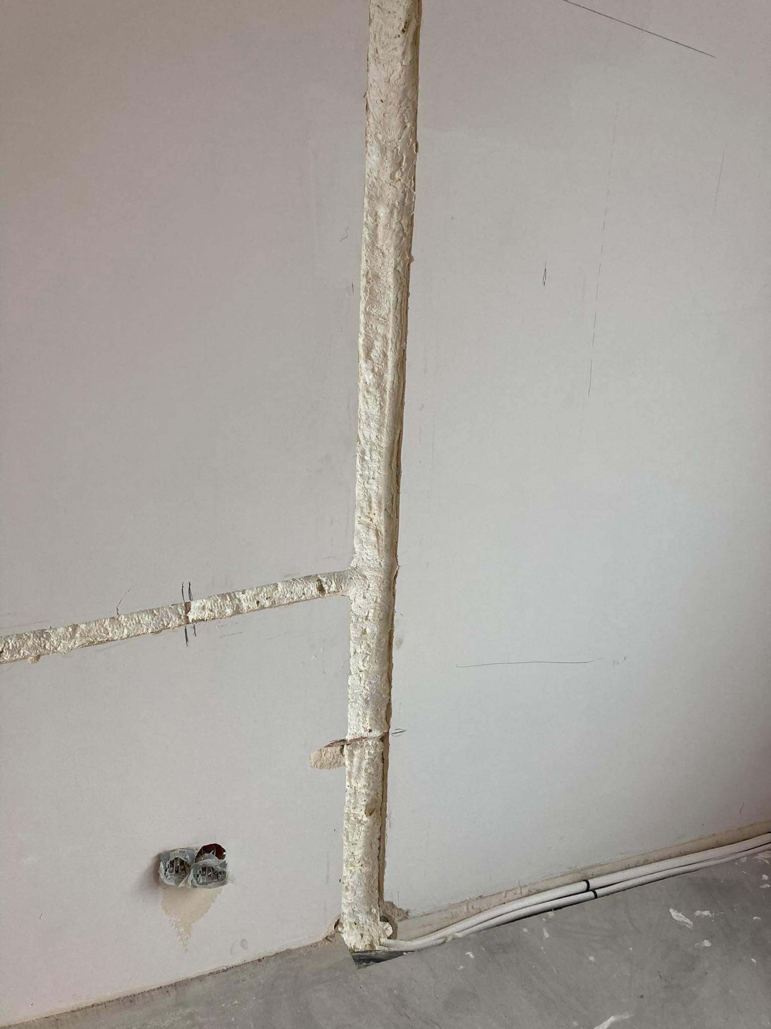 Przygotowanie bruzdy w ścianie pod instalację elektryczną, wypełnionej pianką montażową, widoczne kable elektryczne w podłodze oraz gniazdko elektryczne w ścianie.