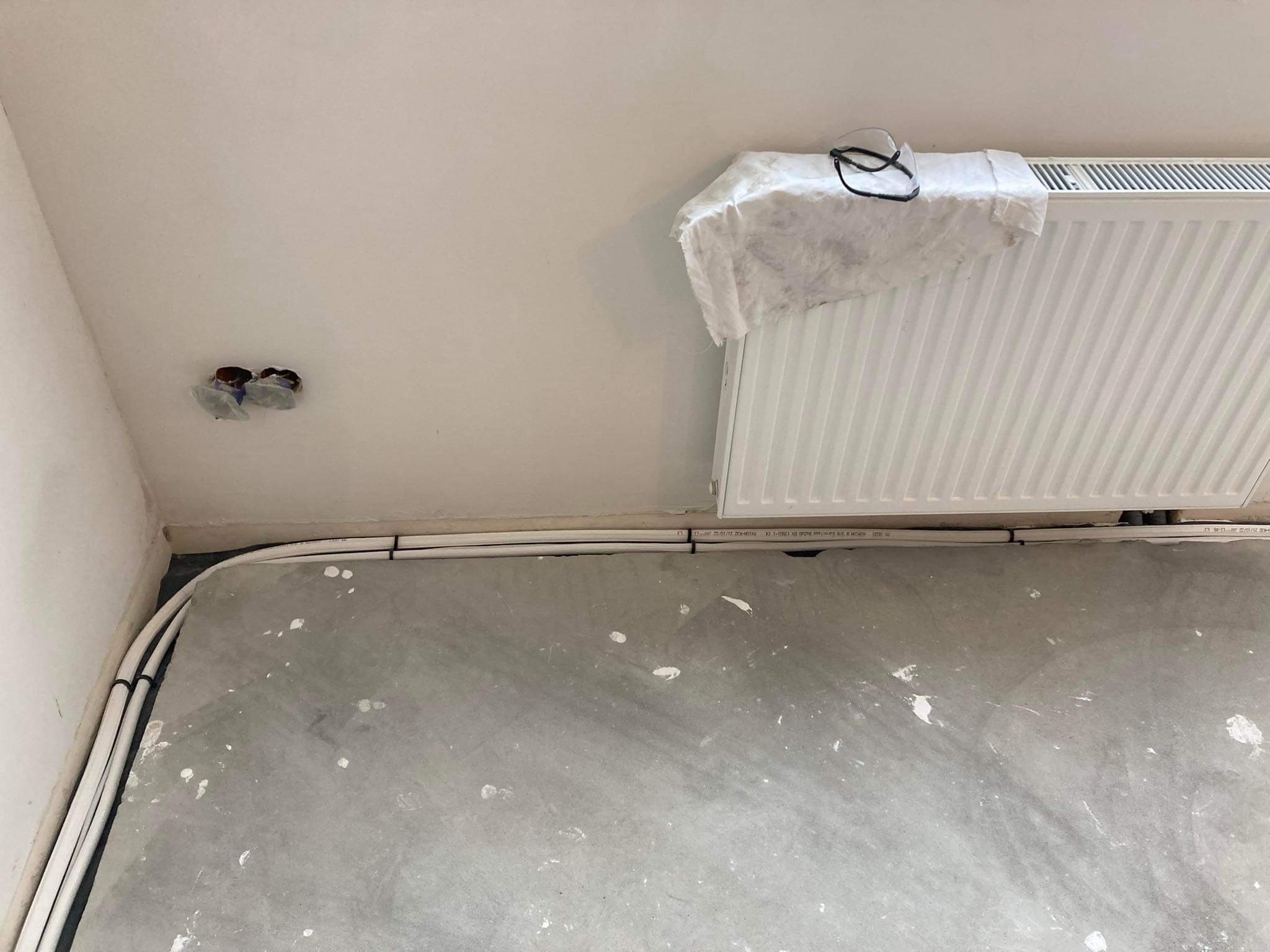 Białe rury grzewcze prowadzone wzdłuż ściany i pod grzejnikiem, zabezpieczonym szmatką i okularami ochronnymi, z widocznymi puszkami elektrycznymi w rogu pomieszczenia w trakcie remontu.