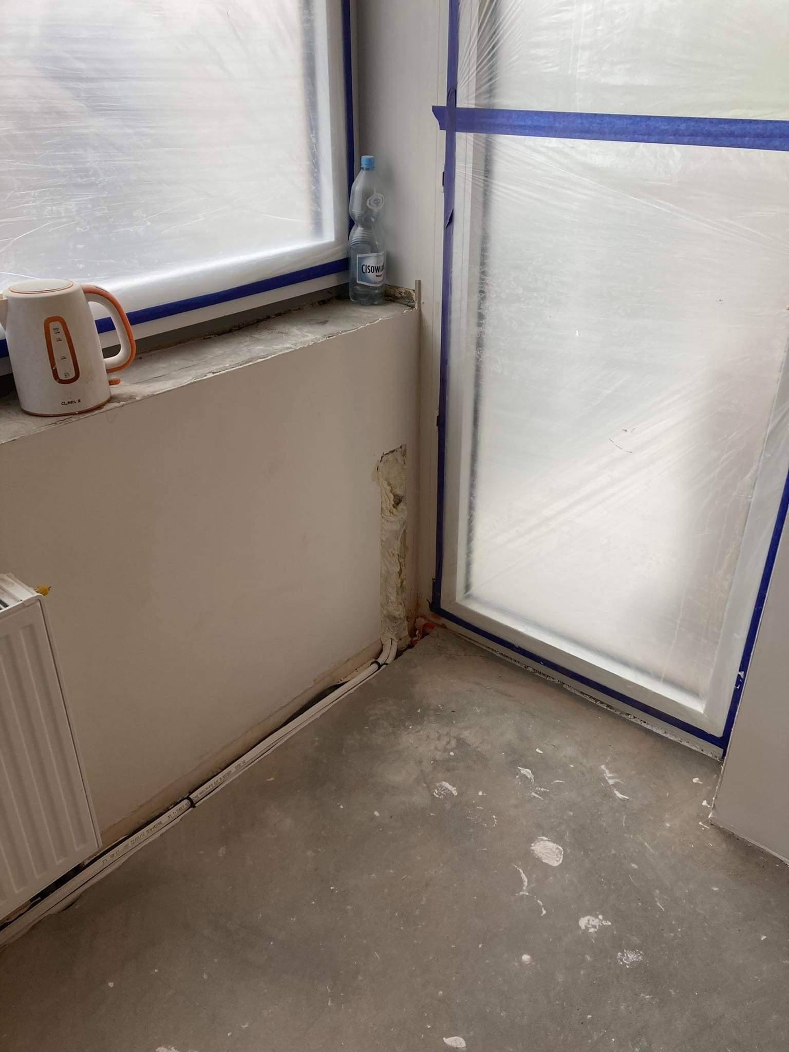 Przygotowania do remontu: okno i ściana zabezpieczone folią malarską przyklejoną niebieską taśmą, widoczne przewody elektryczne w listwie przypodłogowej, czajnik elektryczny i butelka wody...