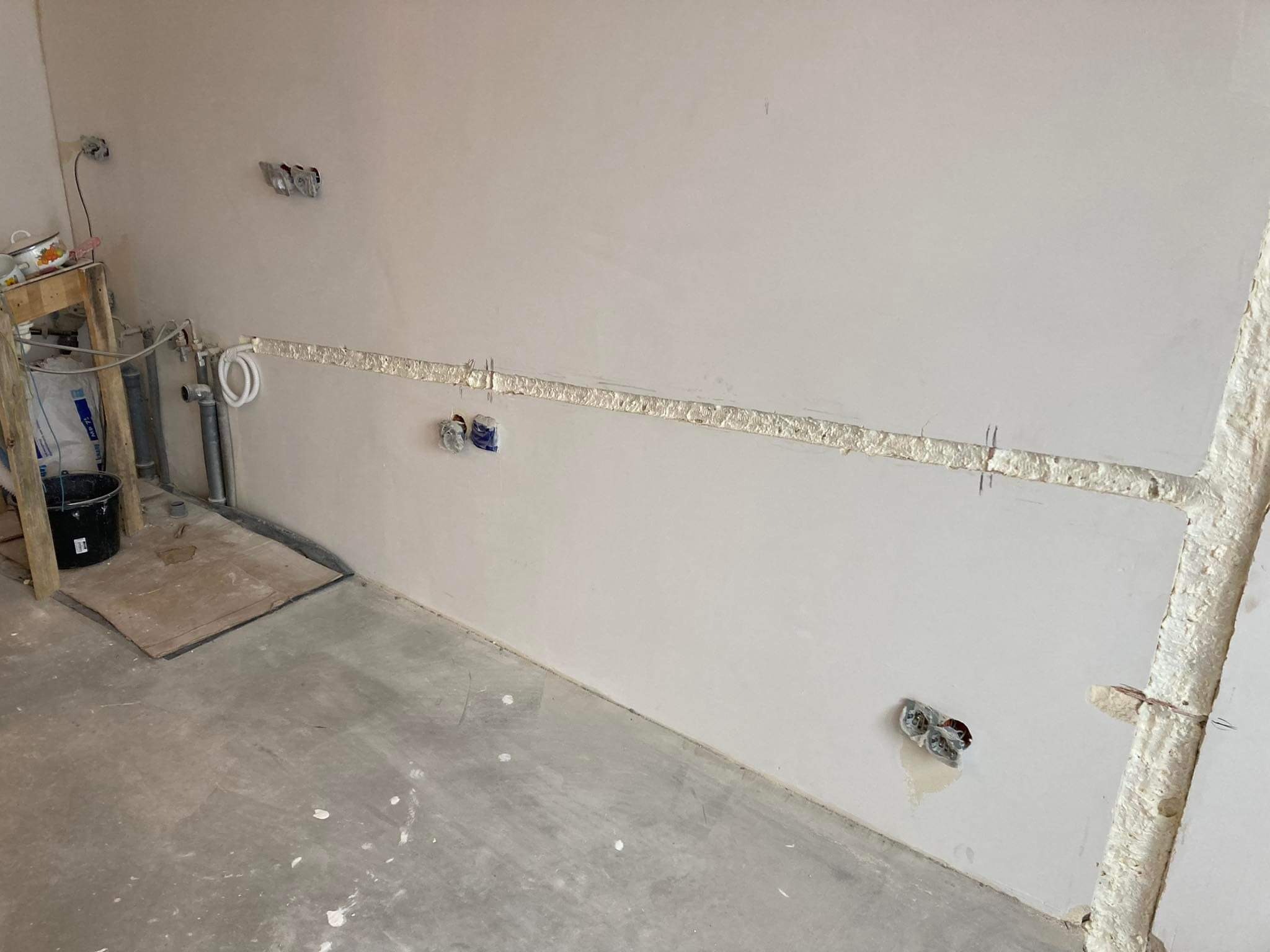 Ściana w trakcie przygotowania do montażu instalacji elektrycznej: wyżłobiony kanał w ścianie, puszki elektryczne zamontowane, widoczne przewody. Surowy beton na podłodze, prowizoryczny stolik...