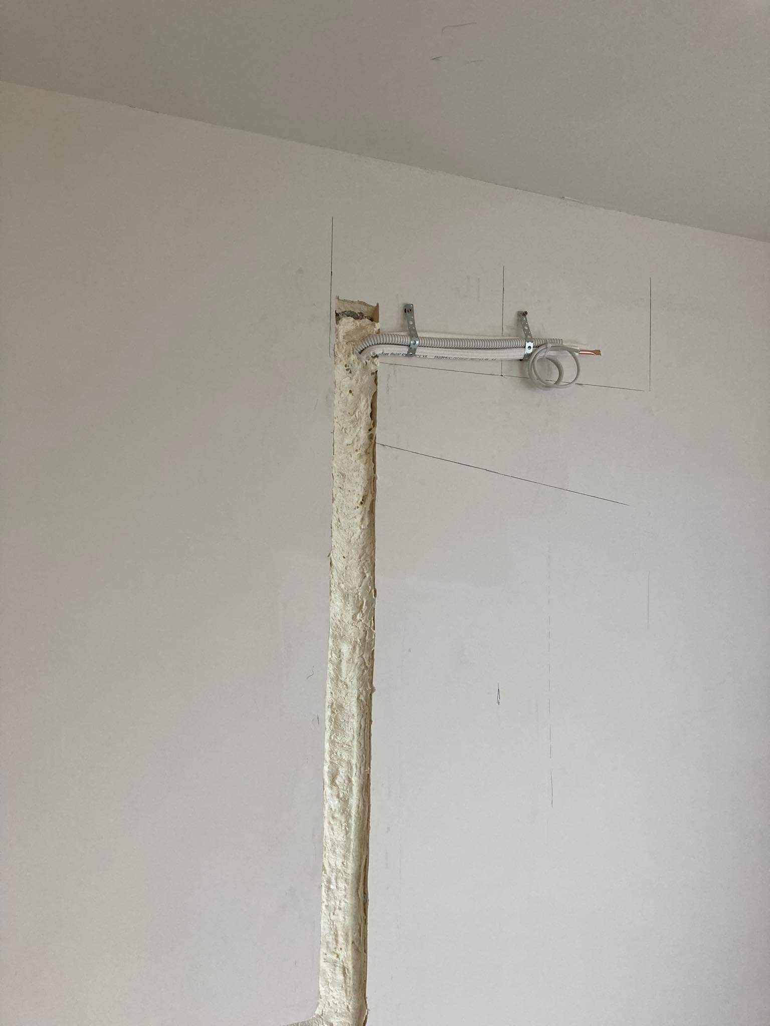 Ściana w trakcie prac instalacyjnych: pionowy wyżłobiony kanał wypełniony pianką montażową, powyżej rurka karbowana przymocowana do ściany metalowymi uchwytami, widoczne przewody elektryczne.