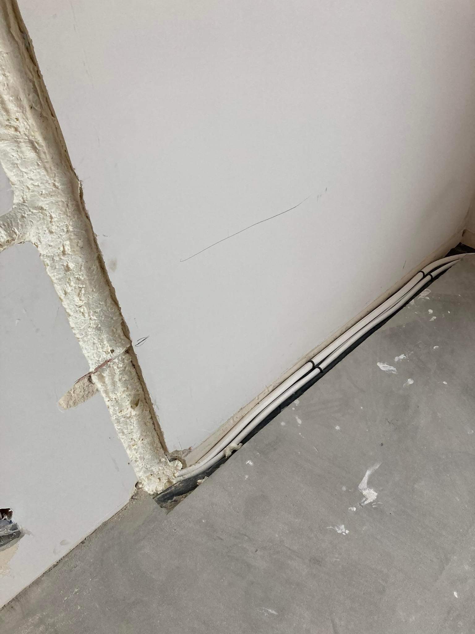 Wewnętrzny róg pomieszczenia z widocznymi przewodami elektrycznymi ułożonymi wzdłuż ściany przy podłodze, zabezpieczonymi pianką montażową w miejscu przejścia przez ścianę. Na podłodze widoczne...