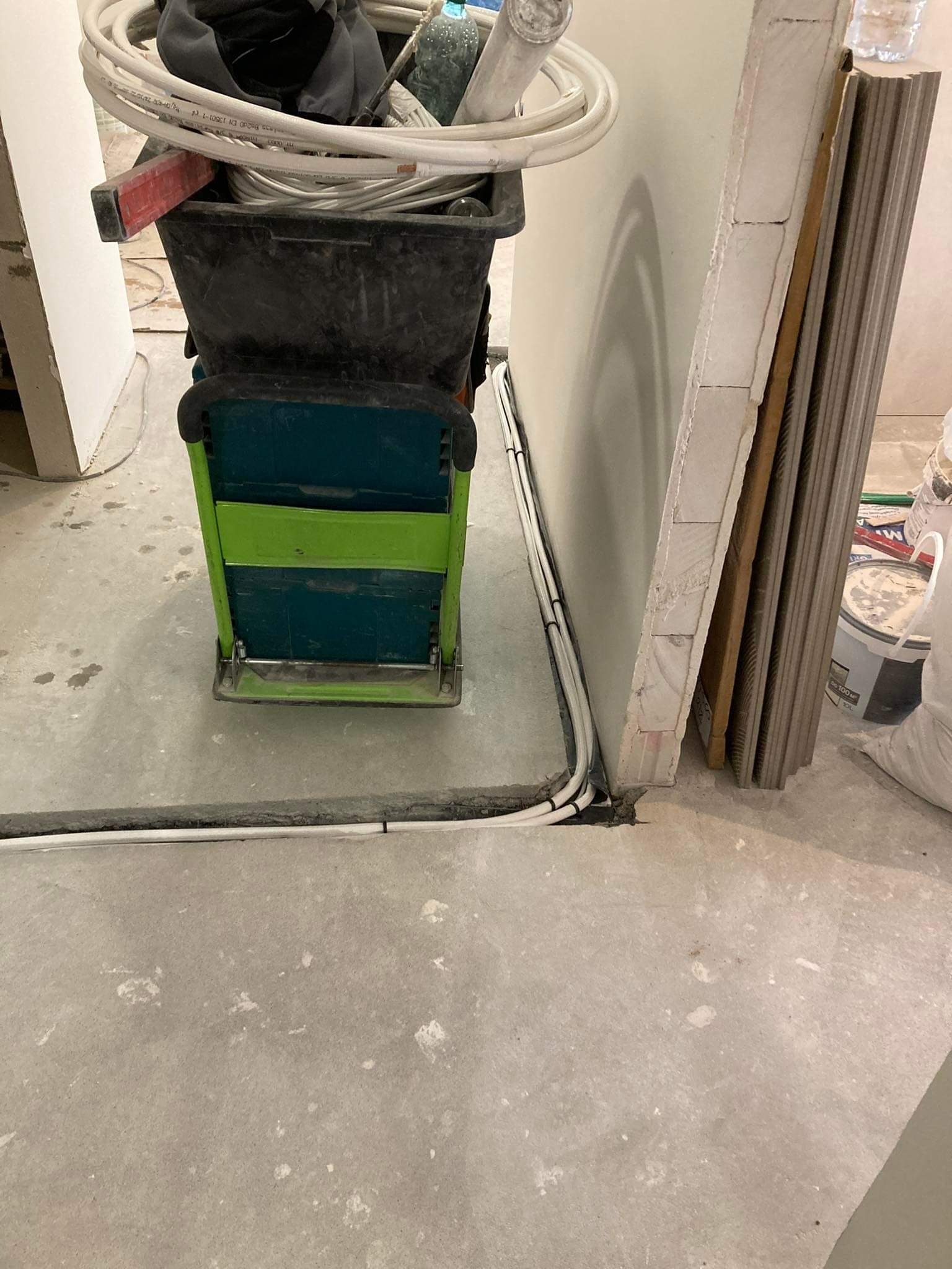Wnętrze w trakcie remontu, widoczne ułożone kable elektryczne wzdłuż ściany, narzędzia w wiadrze na wózku oraz płyty gipsowe oparte o ścianę.