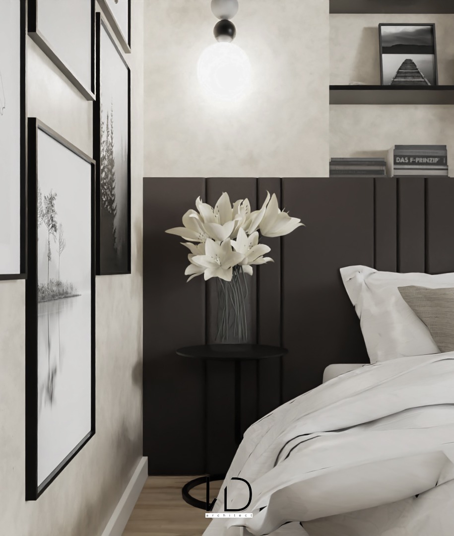 Minimalistyczna sypialnia z czarno-białymi dekoracjami: bukiet białych lilii na czarnym stoliku nocnym, galeria czarno-białych grafik na ścianie, lampa sufitowa i fragment pościeli.